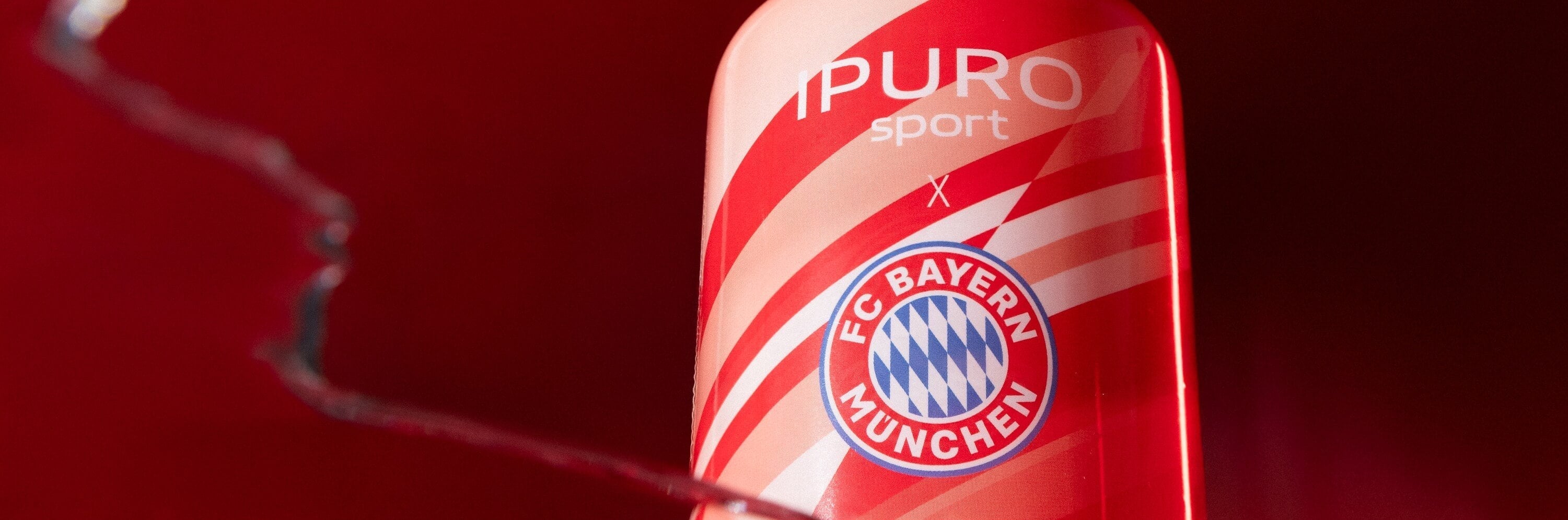 Eine rote Flasche mit diagonalen weißen Streifen und der Aufschrift IPURO sport mit dem Logo des FC Bayern München auf rotem Hintergrund.