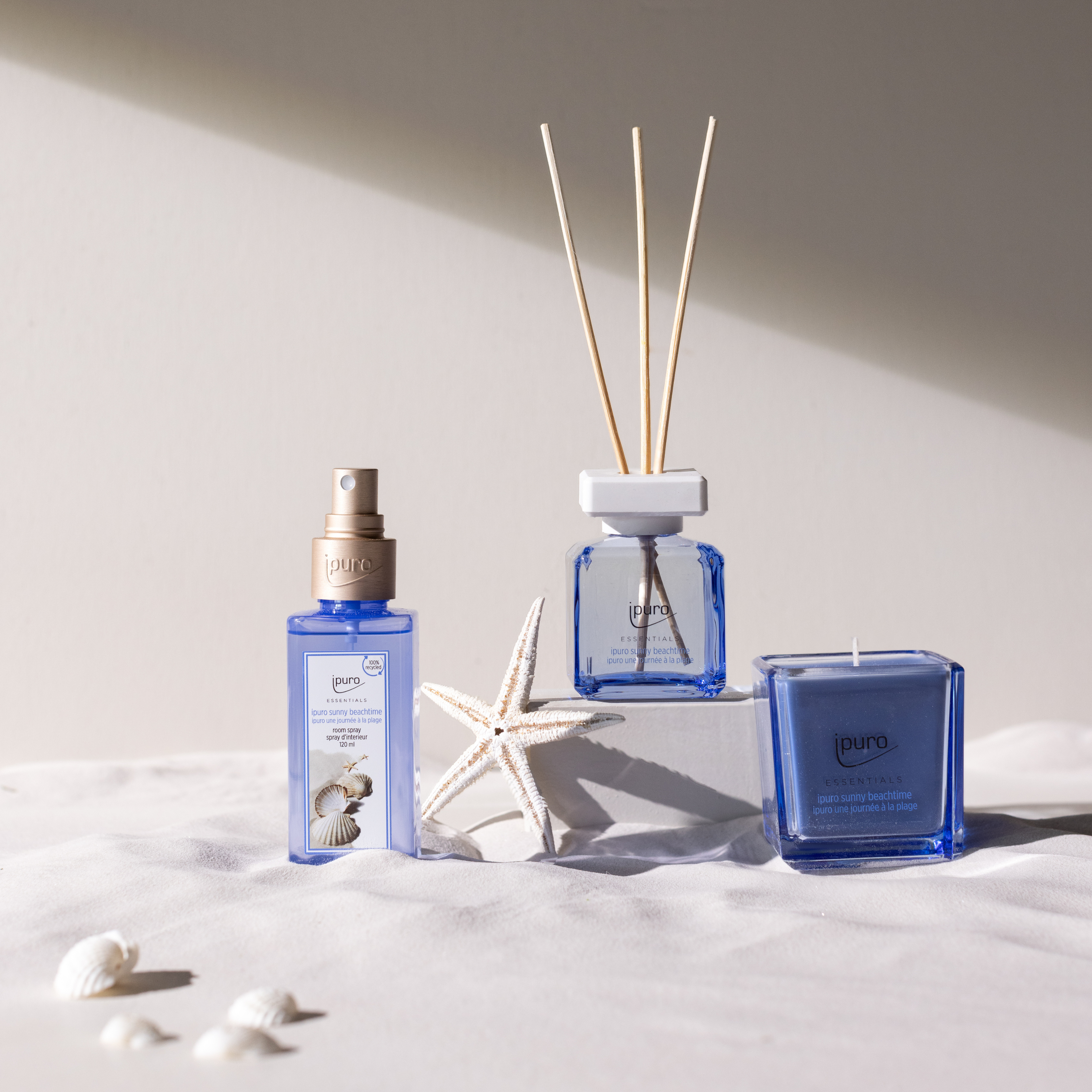 Das ESSENTIALS ipuro sunny beachtime Autoduft von ipuro wird auf einer weißen Fläche mit einem Seestern und Muscheln präsentiert und erinnert an Sommerduft und frische Grapefruit Zitrone Parfum Noten in weichem natürlichem Licht.