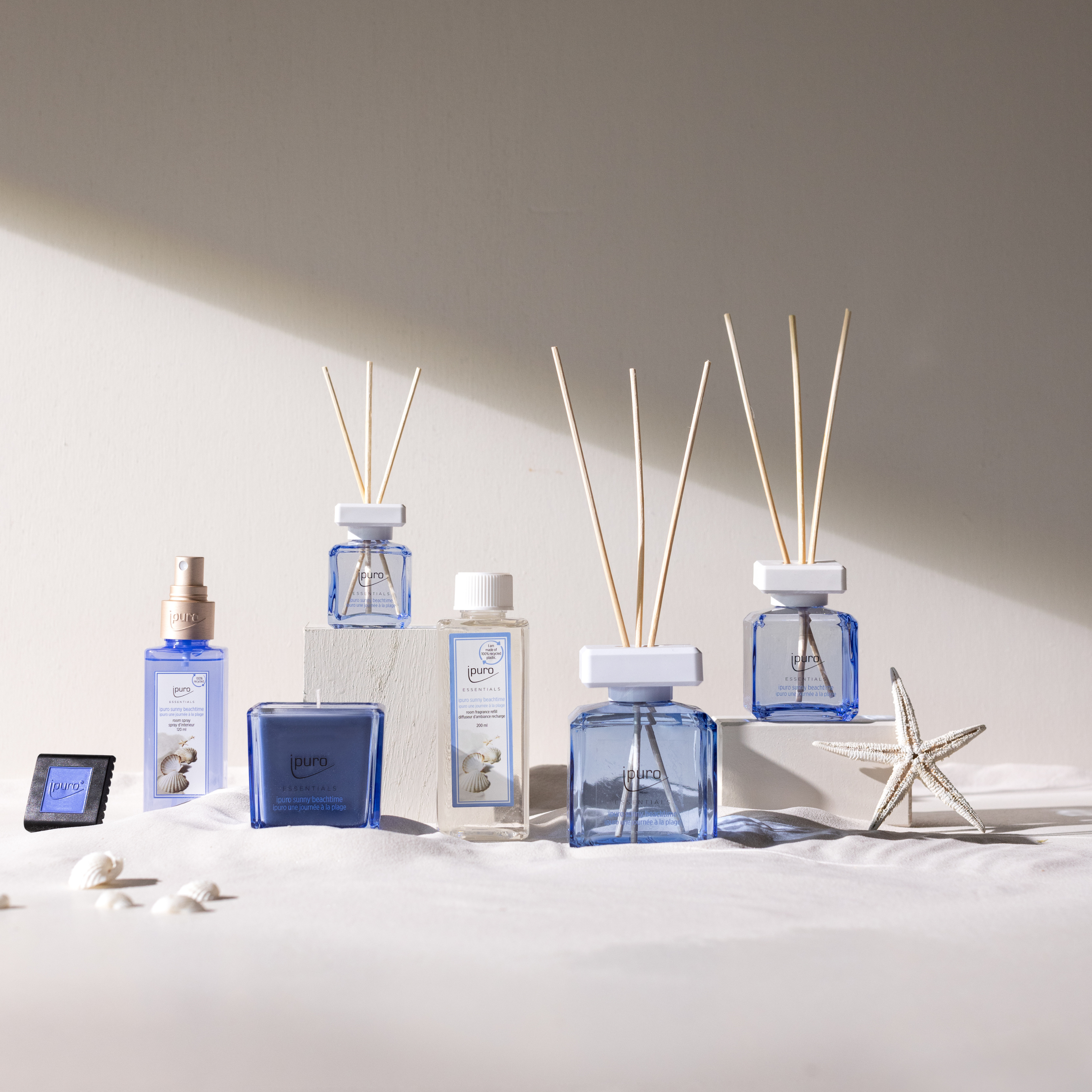 Eine Kollektion von blauen und klaren ipuro ESSENTIALS sunny beachtime Autoduft Diffusoren und Sprühflaschen sind auf einem weißen Tuch mit Muscheln und einem Seestern arrangiert und stehen vor einer neutralen Wand in weichem, natürlichem Licht.