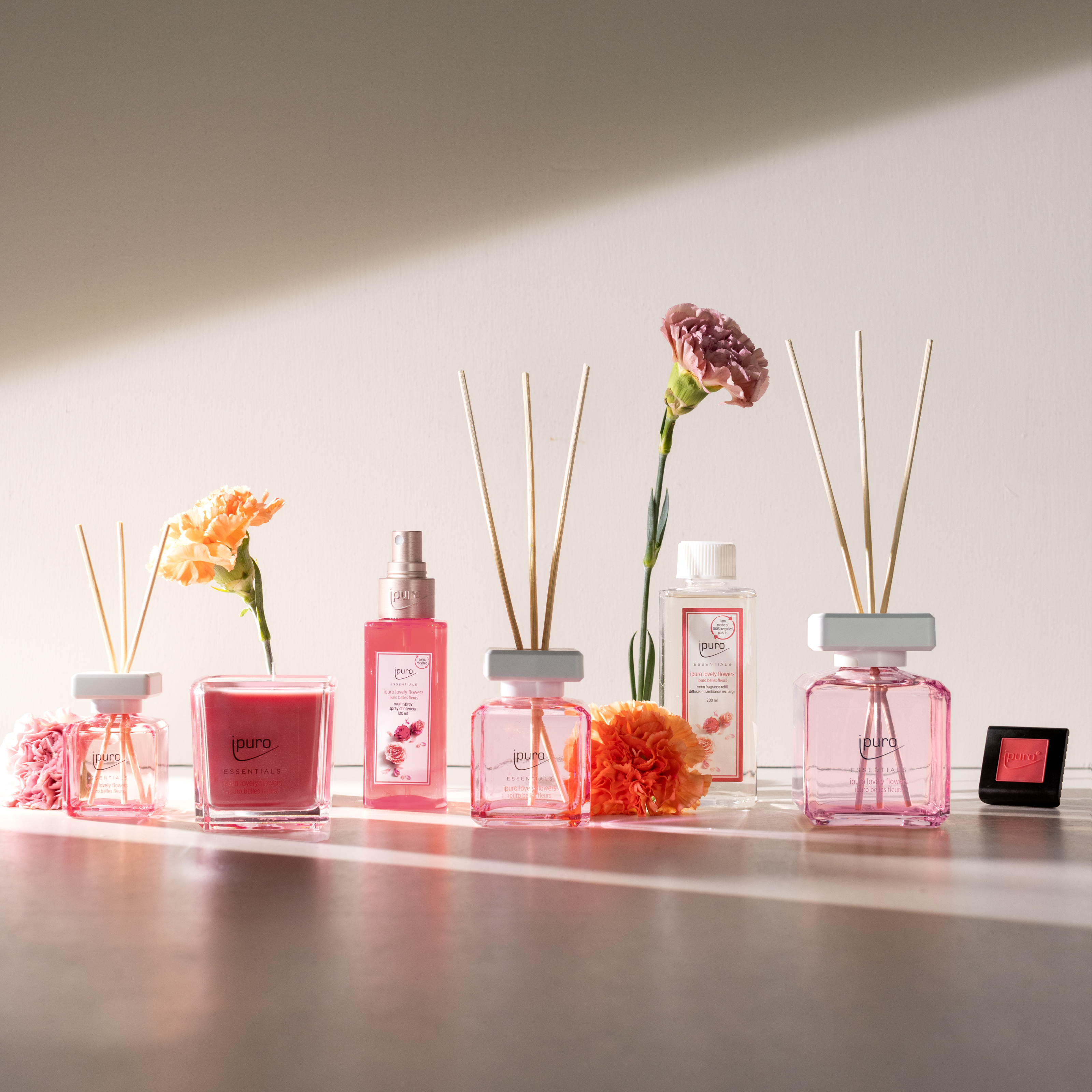 Die ESSENTIALS ipuro lovely flowers Duftkerze von ipuro mit Rosen- und Vanillenoten steht neben rosa Diffusoren, Sprühflaschen und frischen Blumen auf einem sonnendurchfluteten Tisch vor einer weißen Wand.