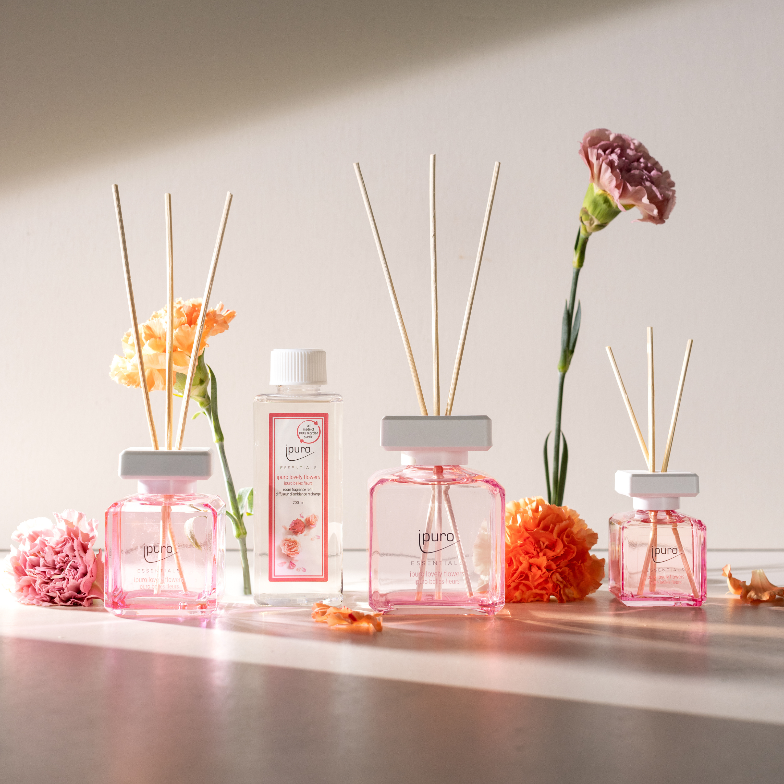 Die ESSENTIALS ipuro lovely flowers Duftkerze von ipuro ist mit mehreren rosafarbenen Diffusoren, einer Nachfüllflasche und orange- und rosafarbenen Blumen auf einer sanft beleuchteten Fläche abgebildet, die sanfte Schatten mit einem Hauch von Rose und Vanille werfen.
