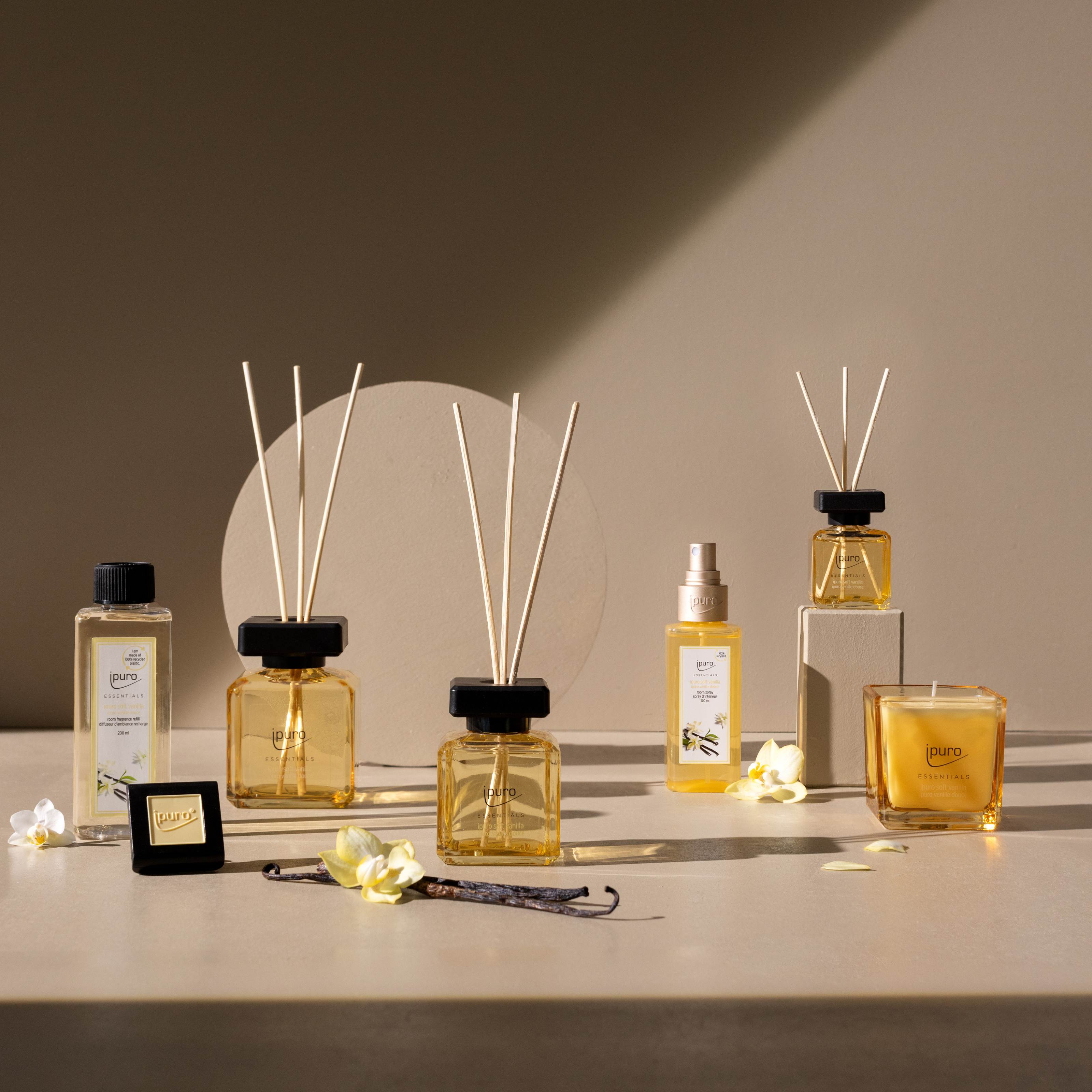 Das ipuro ESSENTIALS soft vanilla Raumduft-Set mit bernsteinfarbenen Diffusoren, einem Raumspray, einer Kerze, einem Seifenstück und einem Flakon wird mit weißen Blütenblättern und Vanilleschoten auf einer neutralen Oberfläche vor einem beigen Hintergrund präsentiert.