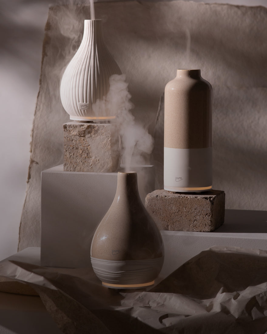 Drei keramische Diffusoren, die Dampf ausstoßen, werden auf strukturierten Steinblöcken und Stoffen ausgestellt, wobei eine weiche, neutrale Beleuchtung ihr minimalistisches, erdiges Design hervorhebt.
