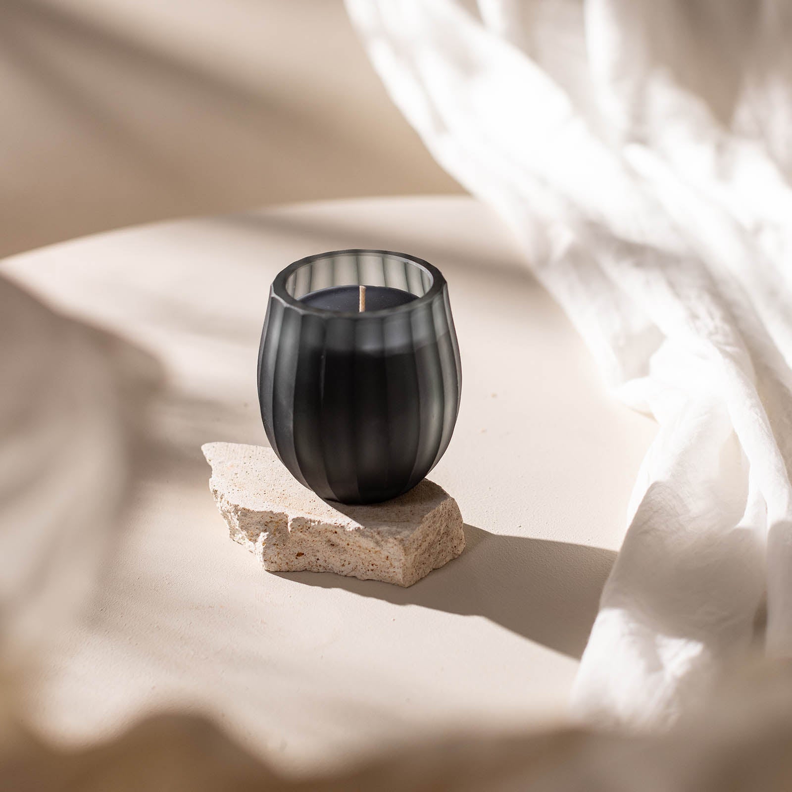 Die Atelier by ipuro gris Duftkerze von ipuro steht in einem schwarzen Milchglashalter auf einer Steinplatte. Weicher weißer Stoff und natürliches Licht unterstreichen ihre elegante Präsenz - perfekt für luxuriöse Kerzenmomente.