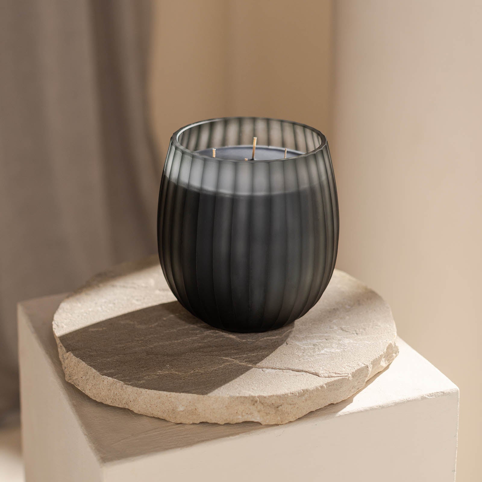 Die Atelier by ipuro gris Duftkerze von ipuro, eine schwarze gerippte Glaskerze mit drei Dochten, ruht auf einer runden Steinplatte auf einem leichten Sockel - perfekt für Fans von luxuriösen Kerzen und minimalistischem Design.