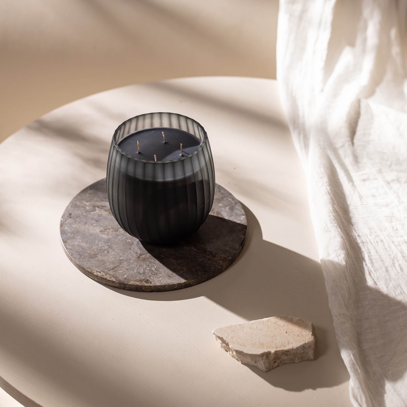 Eine Atelier by ipuro gris Duftkerze aus schwarzem, geripptem Glas mit mehreren Dochten steht auf einem runden Untersetzer aus Stein auf einem hellen Tisch, begleitet von einem rauen Steinfragment und einem schlichten weißen Stoff, der elegant nach rechts drapiert ist.