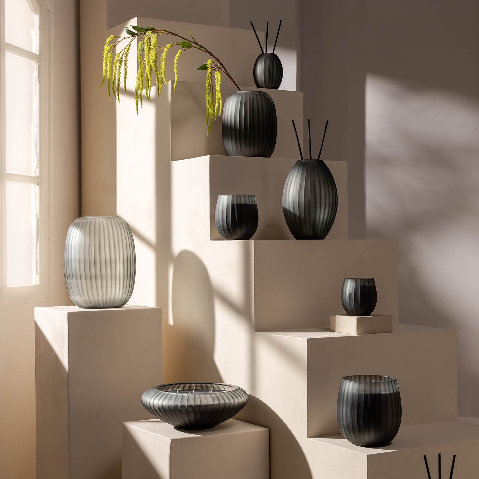 Eine Auslage von gerippten Glasvasen und Schalen in verschiedenen Formen auf cremefarbenen Stufen, die von Sonnenlicht und Grün berührt werden, spiegelt die beruhigenden Sandelholz- und Holznoten von Atelier by ipuro gris Raumduft by ipuro wider.