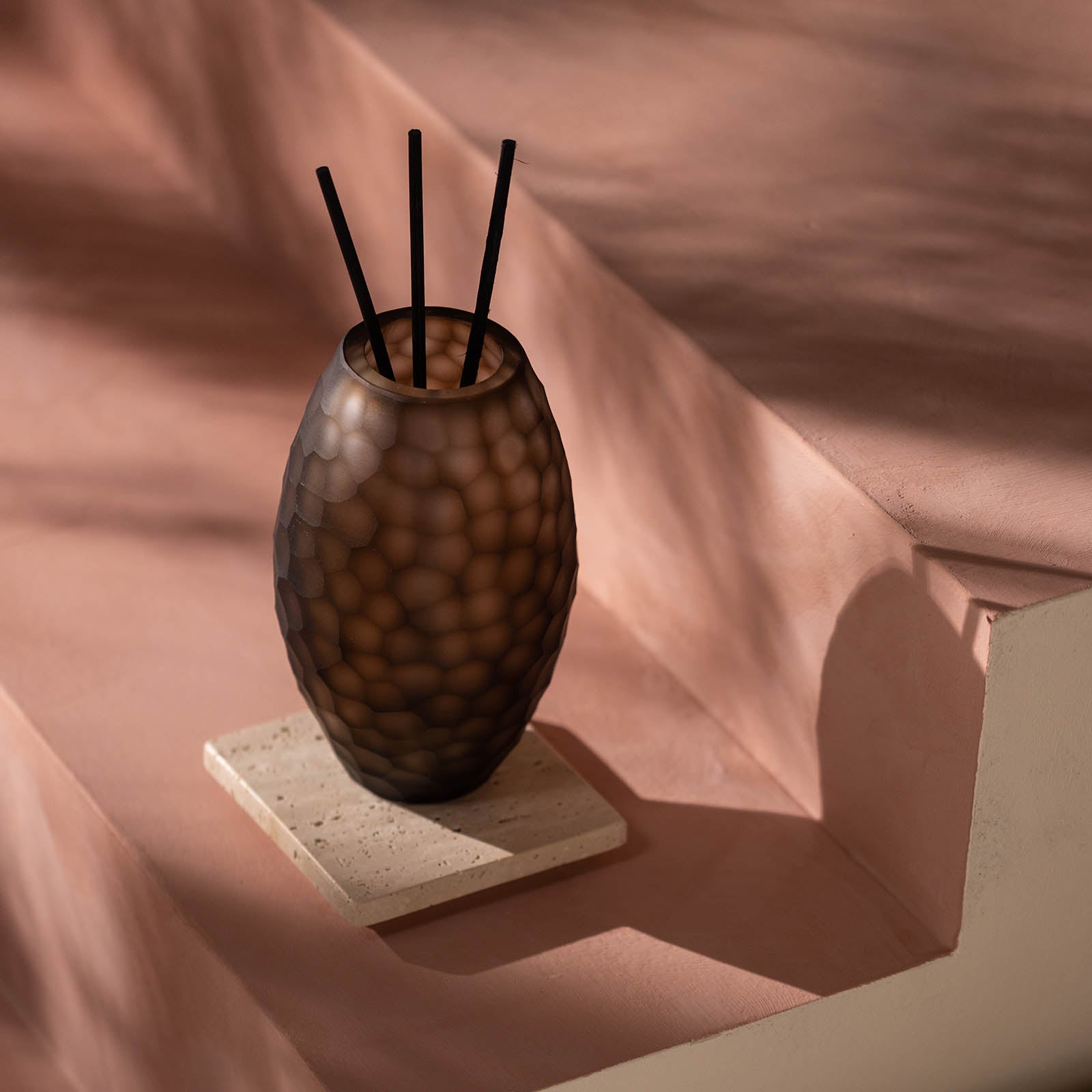 Der Atelier by ipuro brun Diffusor von ipuro mit einem strukturierten braunen Glasdesign mit schwarzen Stäben ruht auf einem hellen Steinuntersetzer auf einer rosa-beigen Stufenfläche in sanften Schatten.
