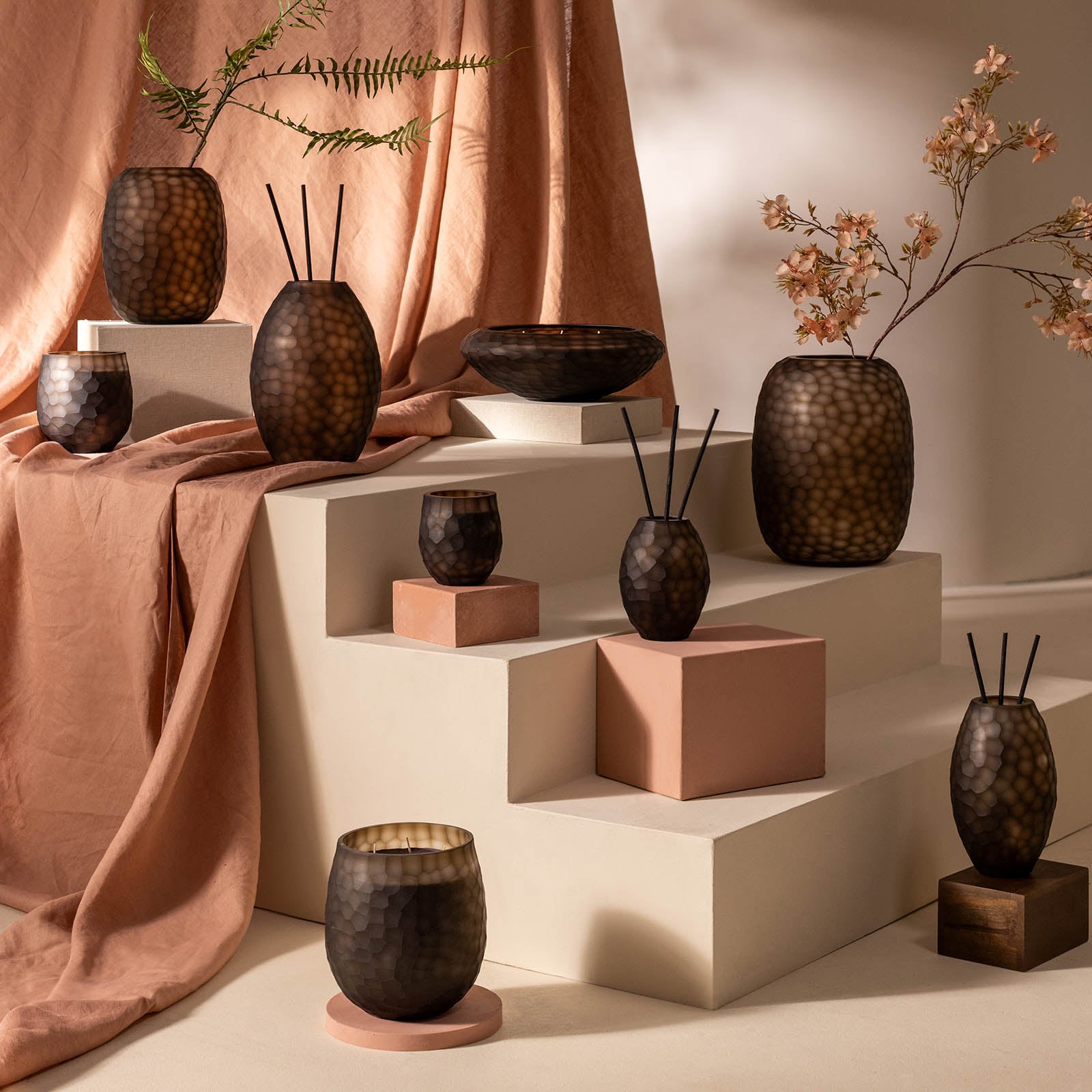 Eine Auslage mit ipuros Atelier by ipuro brun Duftkerze und braunen strukturierten Vasen auf weißen Stufen, mit einem taupefarbenen Tuch und hellen Blumen, schafft ein warmes, modernes dekoratives Ambiente.