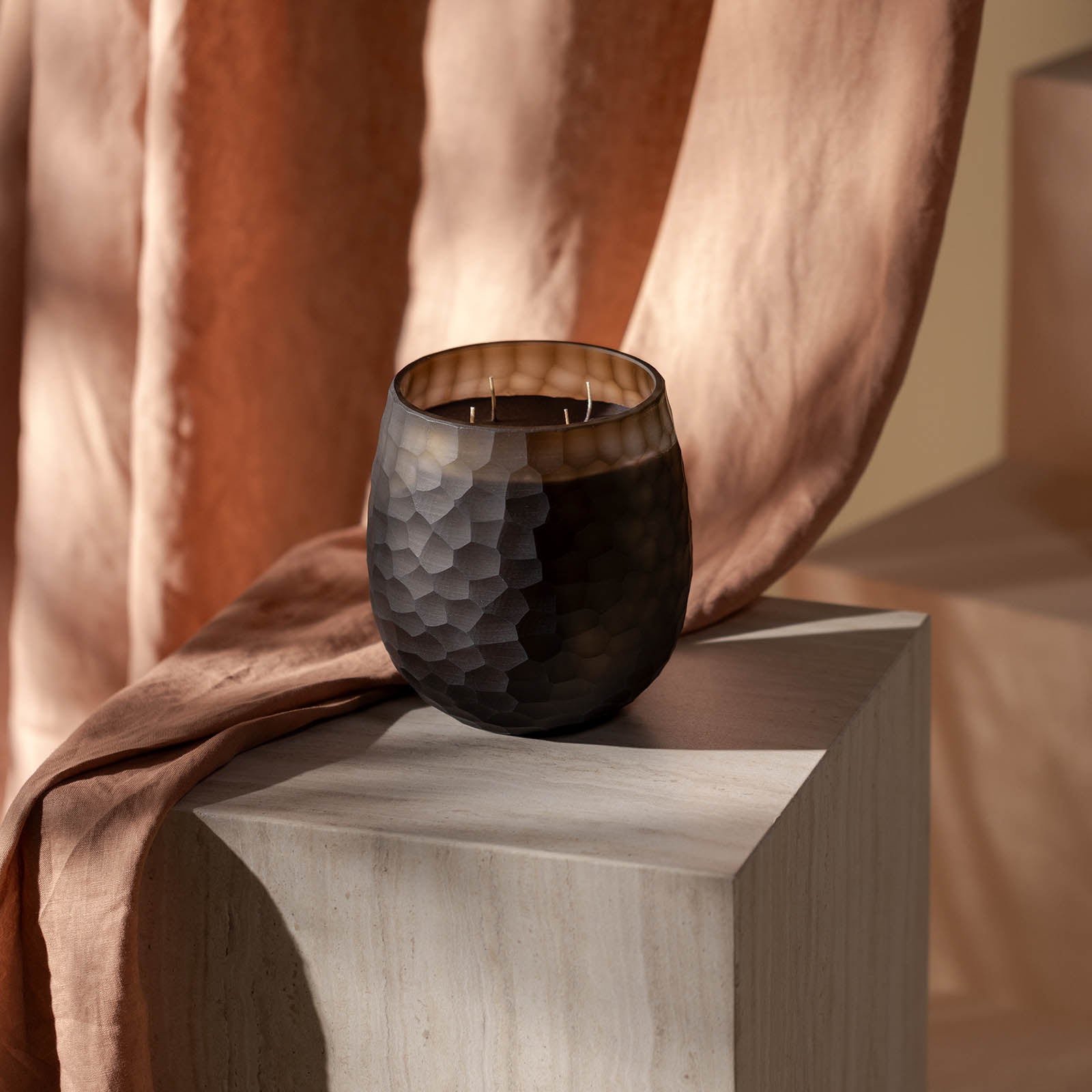 Die Atelier by ipuro brun Duftkerze von ipuro besteht aus einem dunklen, strukturierten Glasgefäß mit drei Dochten, das auf einem hellen, mit pfirsichfarbenem Stoff bespannten Steinsockel steht. Sanftes Licht und neutrale Töne erzeugen eine warme, elegante Stimmung.