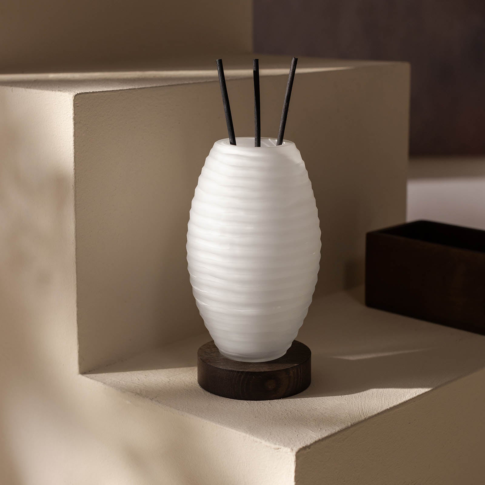 Der ipuro Atelier by ipuro blanc Diffusor, eine weiße gerippte Keramikvase mit schwarzem Schilfrohr, steht auf einem dunklen Holzsockel auf einer beigen Stufenfläche in sanftem Tageslicht.