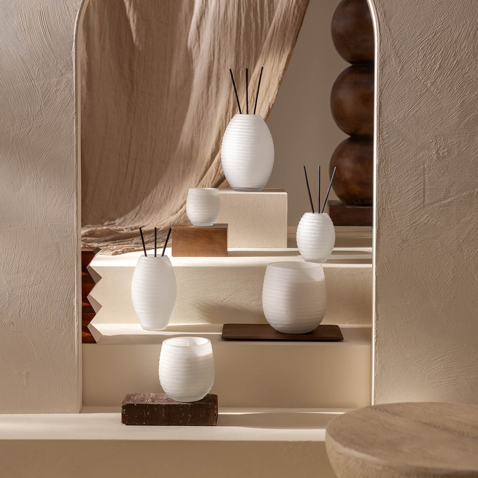 Der Atelier by ipuro blanc Diffusor von ipuro wird auf neutral getönten Stufen mit hölzernen Akzenten, strukturierten Wänden und beigefarbenem, drapiertem Stoff in einem minimalistischen, erdigen Ambiente präsentiert.