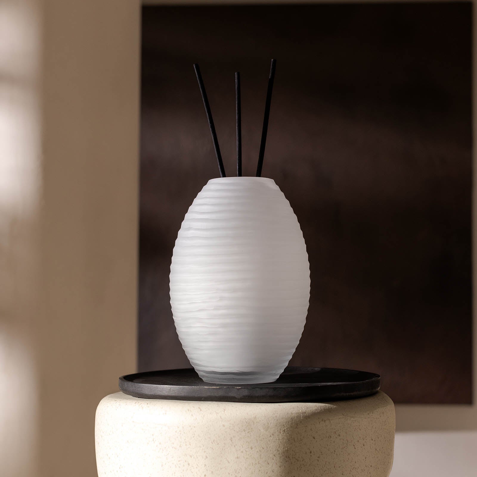 Der Atelier by ipuro blanc Diffusor mit einer Vase aus mattiertem, geripptem Glas und drei schwarzen Stäben steht auf einem runden, schwarzen Tablett auf einem beigen, strukturierten Sockel vor einem dunklen, rechteckigen Hintergrund.