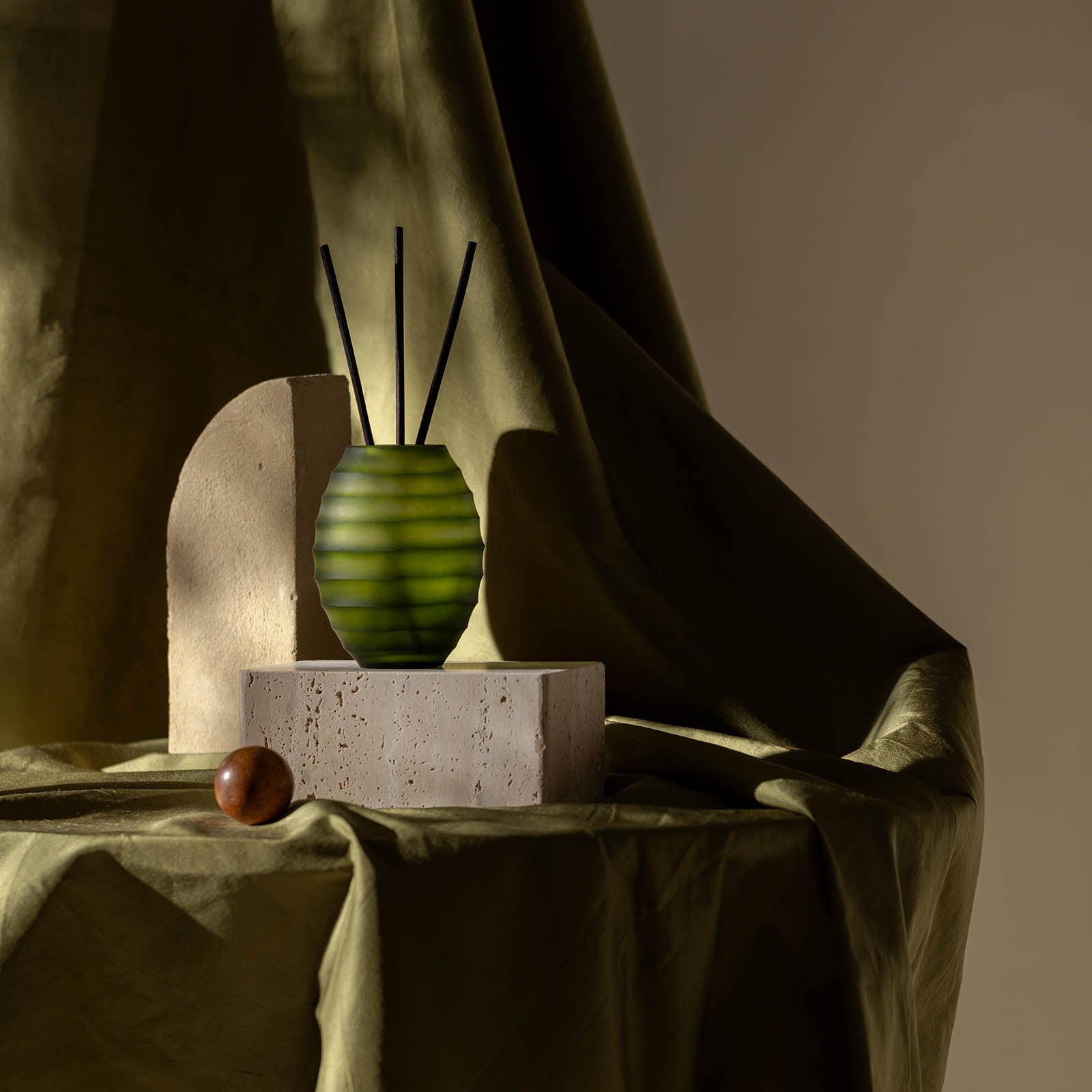 Ein Atelier by ipuro vert Diffusor mit schwarzem Stäbchen von ipuro ruht auf einem Steinblock neben einem geschwungenen beigen Objekt und einer braunen Kugel, die auf einem drapierten olivgrünen Stoff angeordnet sind und für auffällige Licht- und Schatteneffekte sorgen.