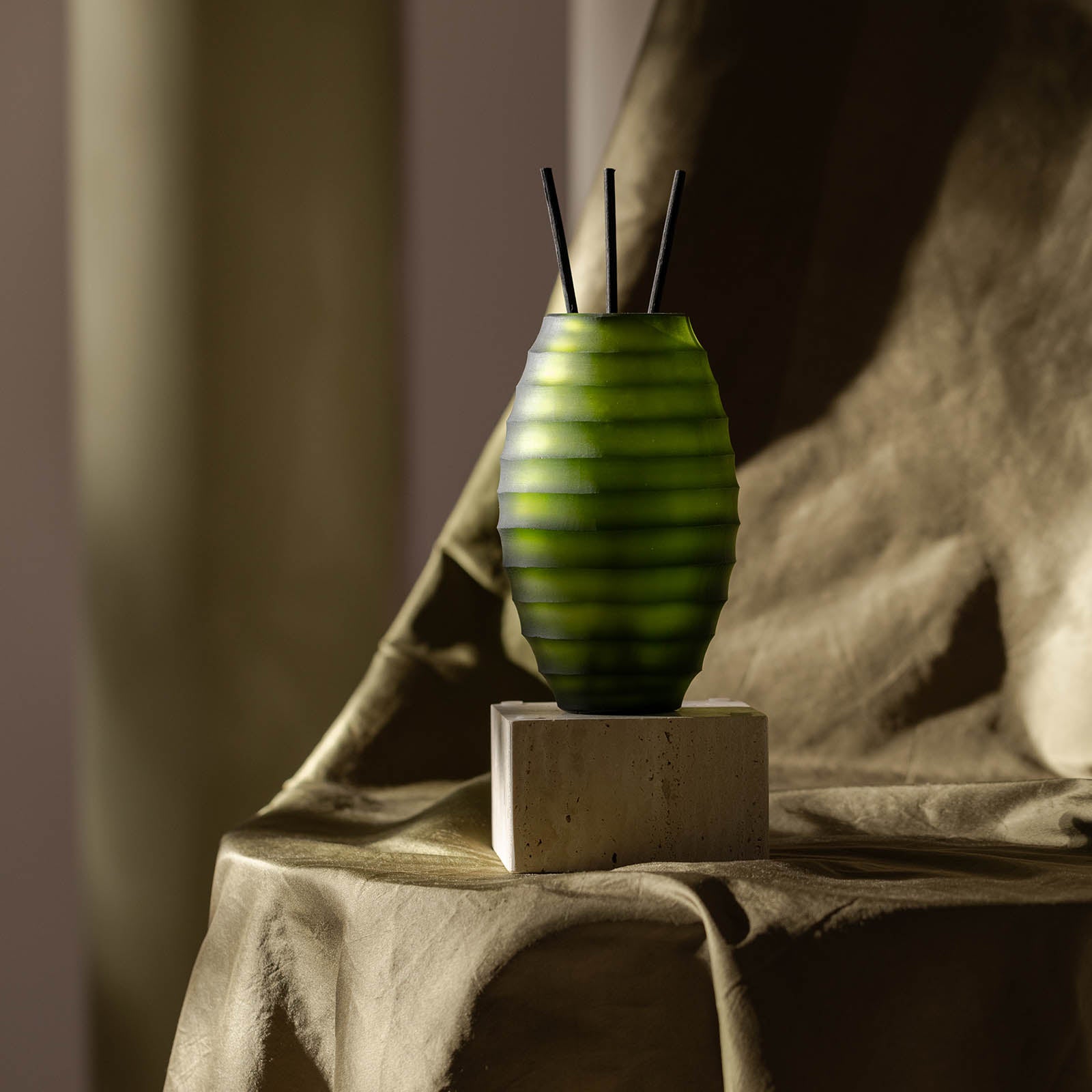 Der ipuro Atelier by ipuro vert Diffusor, eine grüne, gerippte Vase mit drei schwarzen Schilfrohren, ruht auf einem kleinen Steinblock, der mit beigefarbenem, strukturiertem Stoff überzogen ist und von sanftem natürlichem Licht beleuchtet wird.