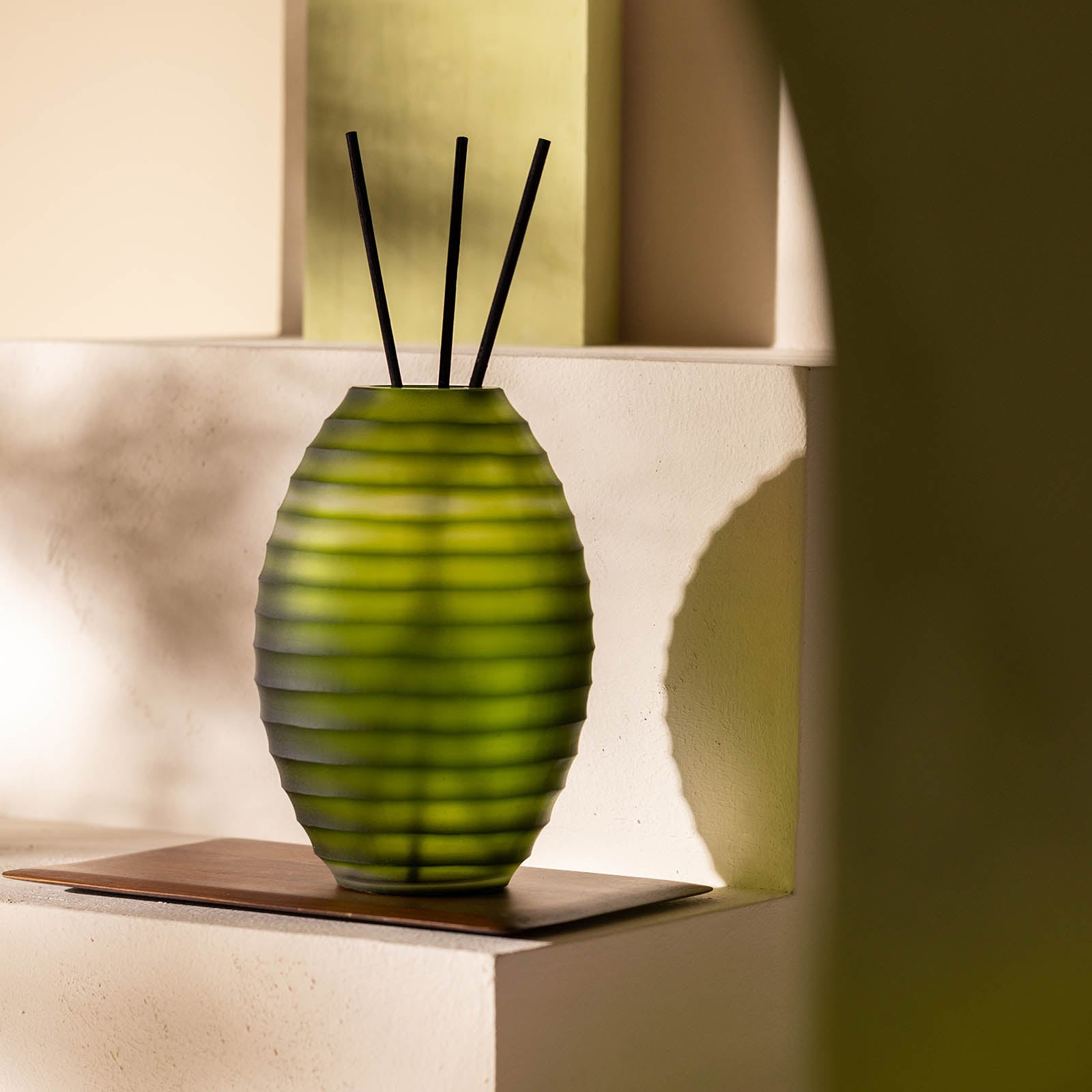 Der Atelier by ipuro vert Diffusor von ipuro hat ein grünes, geripptes Glasdesign mit drei schwarzen Schilfrohren, die auf einem Holztablett auf einer weißen Oberfläche präsentiert werden - sein eleganter Stil ist ideal für die Präsentation luxuriöser Düfte.