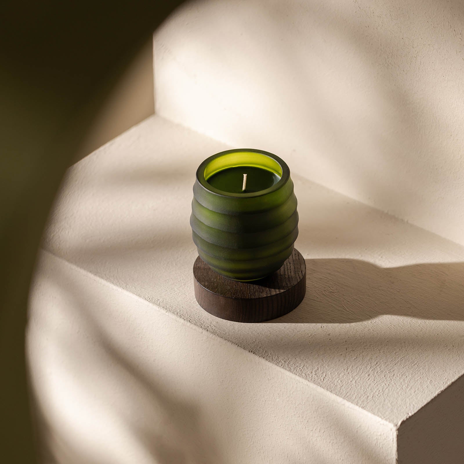 Eine grüne gerippte Atelier by ipuro vert Duftkerze von ipuro ruht auf einem runden dunklen Holzuntersetzer auf einer hellen strukturierten Oberfläche. Sonnenlicht und Schatten schaffen ein beruhigendes Ambiente - ideal für Fans von Atelier by ipuro Duftkerzen.