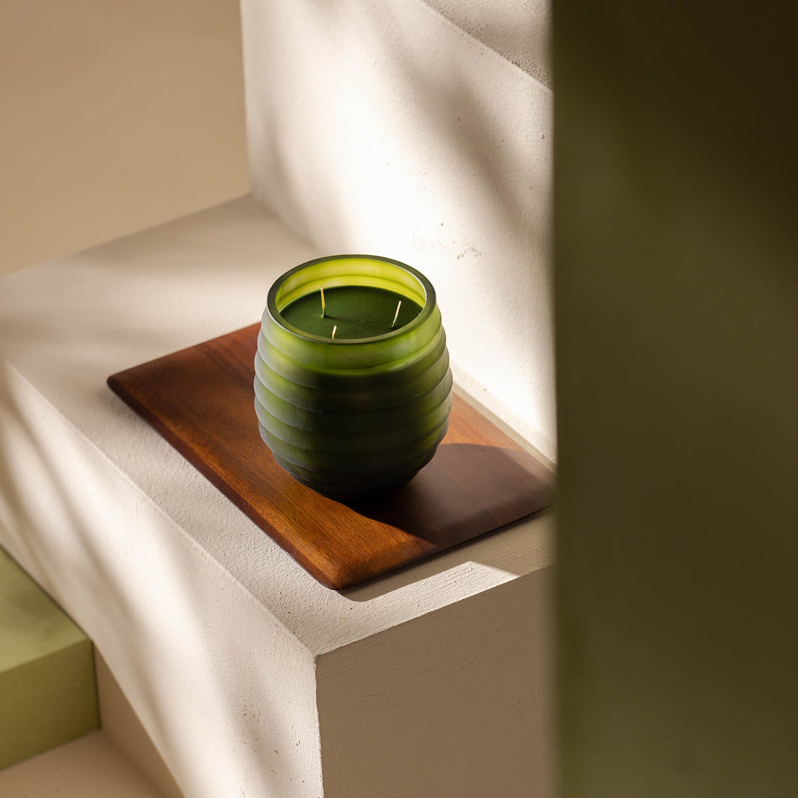 Die Atelier by ipuro vert Duftkerze by ipuro, eine grüne gerippte Glaskerze mit zwei Dochten, ruht auf einem Holztablett auf weißen minimalistischen Stufen. Das Sonnenlicht wirft sanfte Schatten und schafft eine ruhige und gemütliche Atmosphäre.