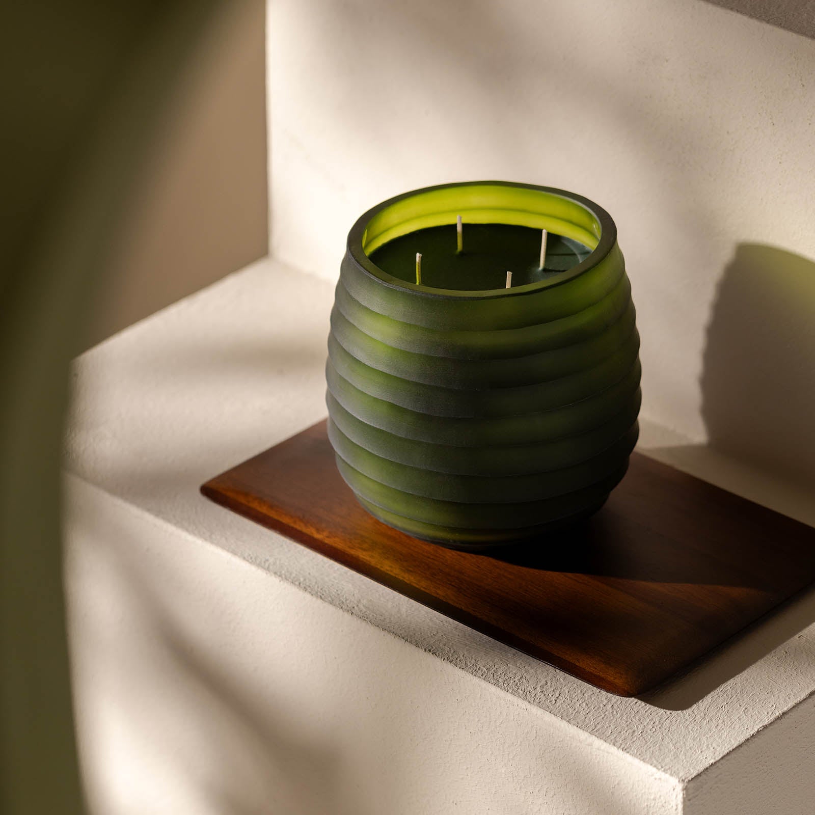 Die Atelier by ipuro vert Duftkerze von ipuro, eine grüne gerippte Glaskerze mit mehreren Dochten, ruht auf einem Holztablett auf einer leicht gestuften, in natürliches Licht getauchten Fläche, ideal für Fans von handgefertigten Kerzen.