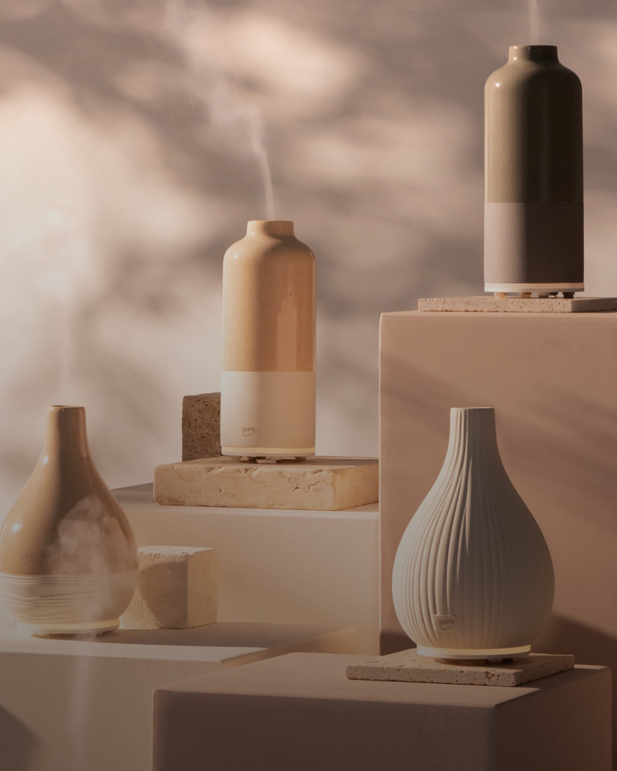 Drei keramische Diffusoren in neutralen Tönen geben sichtbare Nebelströme ab, die auf gestapelten beigen Blöcken mit einem weichen, verschwommenen Hintergrund angeordnet sind. Das Design ist minimalistisch und modern, mit verschiedenen Formen und Texturen.