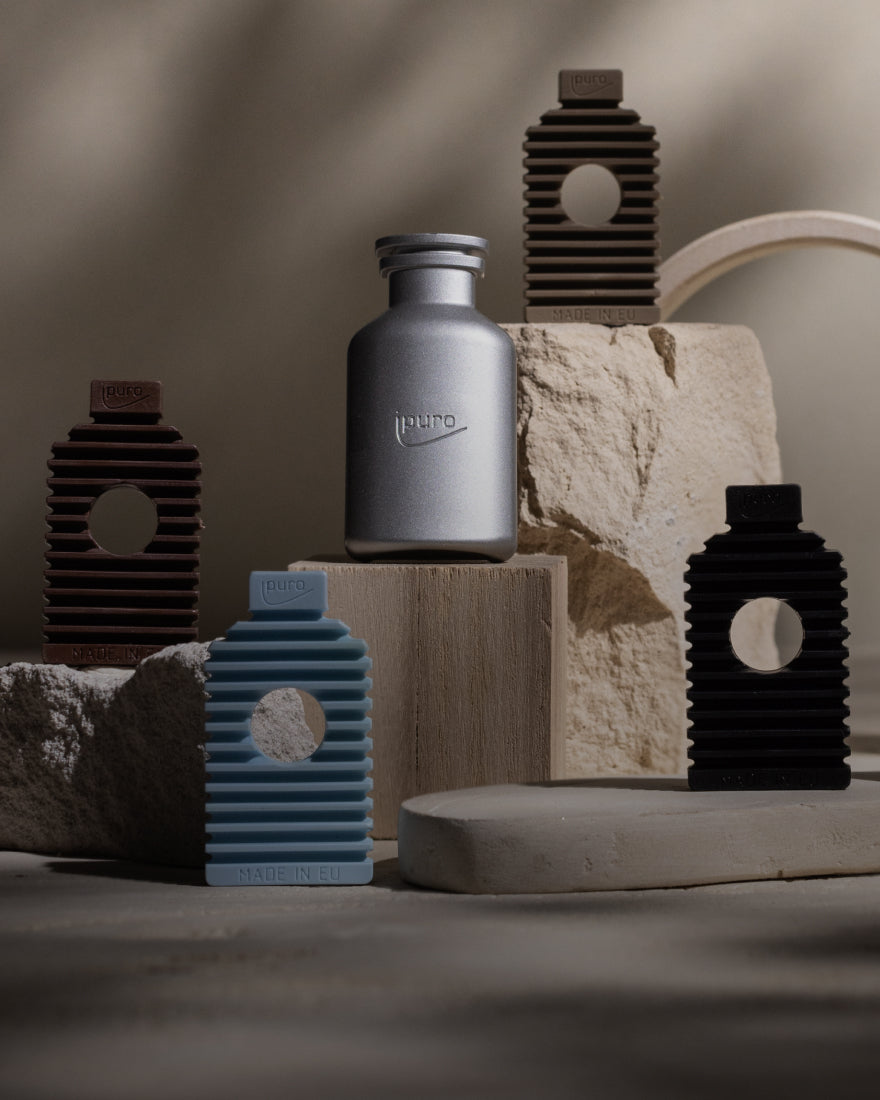 Eine Metallflasche und fünf gerippte, rechteckige Halterungen in Blau, Schwarz und Braun sind auf strukturierten beigen Steinen angeordnet, mit weichen Schatten und neutraler Beleuchtung im Hintergrund.