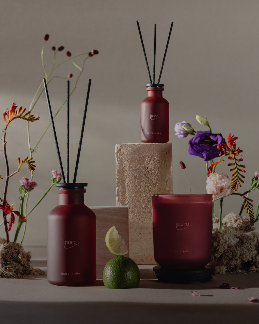 Drei burgunderfarbene Duftprodukte, darunter zwei Diffusoren mit schwarzen Stäbchen und eine Kerze, sind zusammen mit einem Limettenkeil, Moos und bunten Blumen auf einem Steinsockel vor einem neutralen Hintergrund arrangiert.