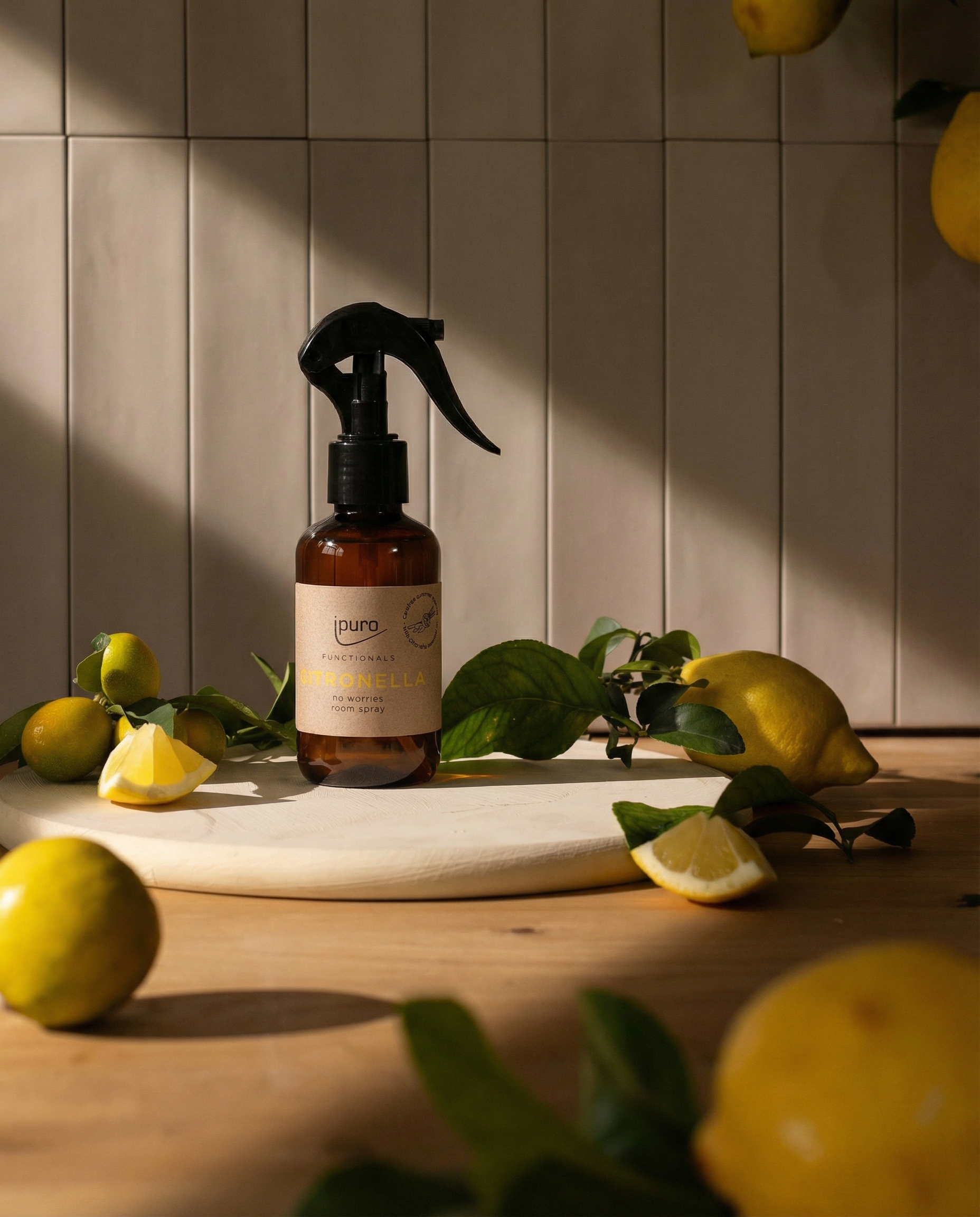 Eine braune FUNCTIONALS ipuro citronella Raumspray 150ml Flasche von ipuro steht auf einer runden weißen Tafel, umgeben von frischen Zitronen, grünen Blättern und Zitronengras auf einer Holzfläche mit gefliestem Wandhintergrund und natürlichem Sonnenlicht.