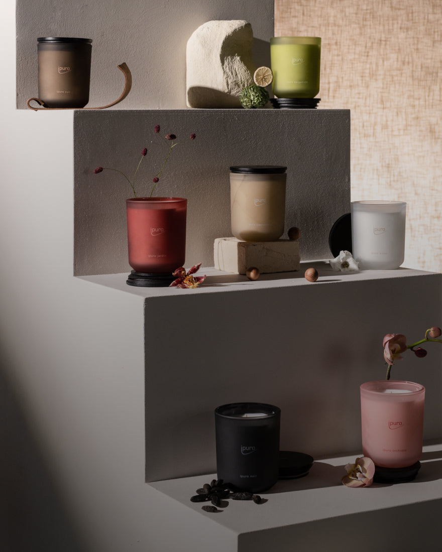 Ein Arrangement aus sieben Kerzen in verschiedenfarbigen Gläsern, die auf gestaffelten weißen Regalen stehen und von dekorativen Gegenständen wie Blumen, Steinen und Fruchtscheiben umgeben sind.