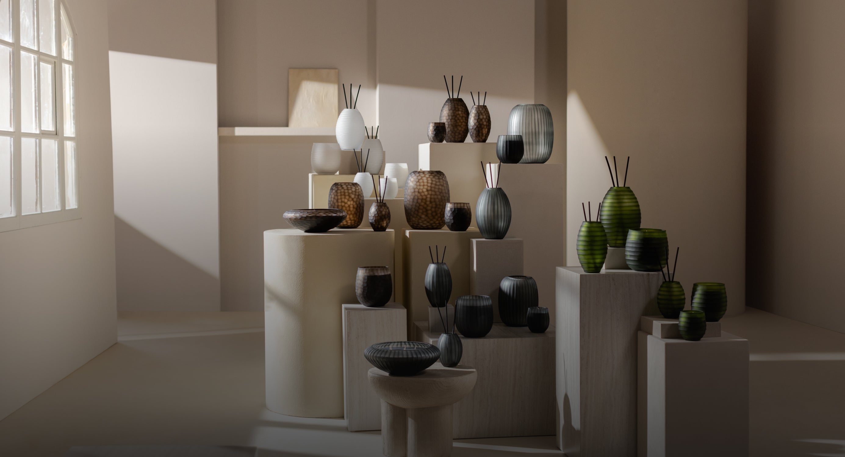 Eine Sammlung von dekorativen Vasen und Schalen in verschiedenen Größen und Farben, ausgestellt auf Sockeln in einem sanft beleuchteten, minimalistischen Raum mit Licht, das durch ein großes Fenster fällt.