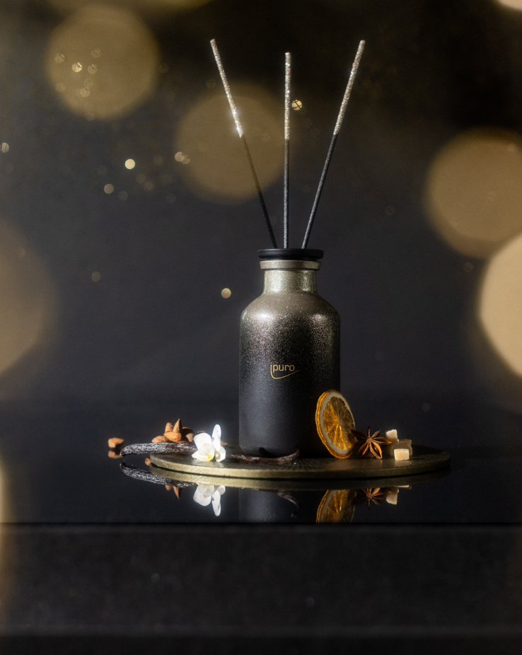 Der LIMITED EDITION ipuro warm glamour Raumduft 240 ml von ipuro steht mit Schilfrohrstäbchen auf einem runden Tablett, umgeben von getrockneten Orangenscheiben, Vanilleschoten und Sternanis - ein gemütlicher Winterduft vor einem dunklen, goldenen Hintergrund.