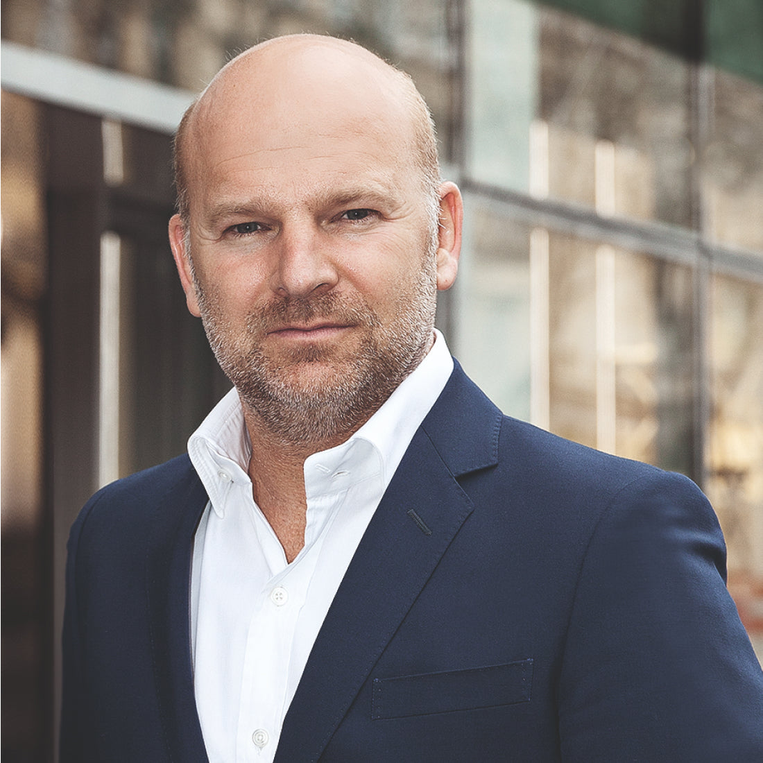Ein Mann mit rasiertem Kopf und kurzem Bart, der ein weißes Hemd und einen dunklen Blazer trägt, steht draußen vor einem modernen Glasgebäude und schaut selbstbewusst in die Kamera.