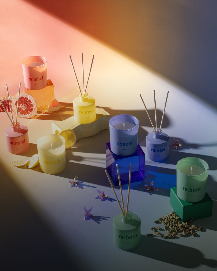 Bunte Kerzen und Rohrdiffusoren mit Wörtern wie re:mind, re:fresh, re:new und re:born sind mit Fruchtscheiben, Schmetterlingen und Gewürzen unter einem Farbverlaufslicht arrangiert.