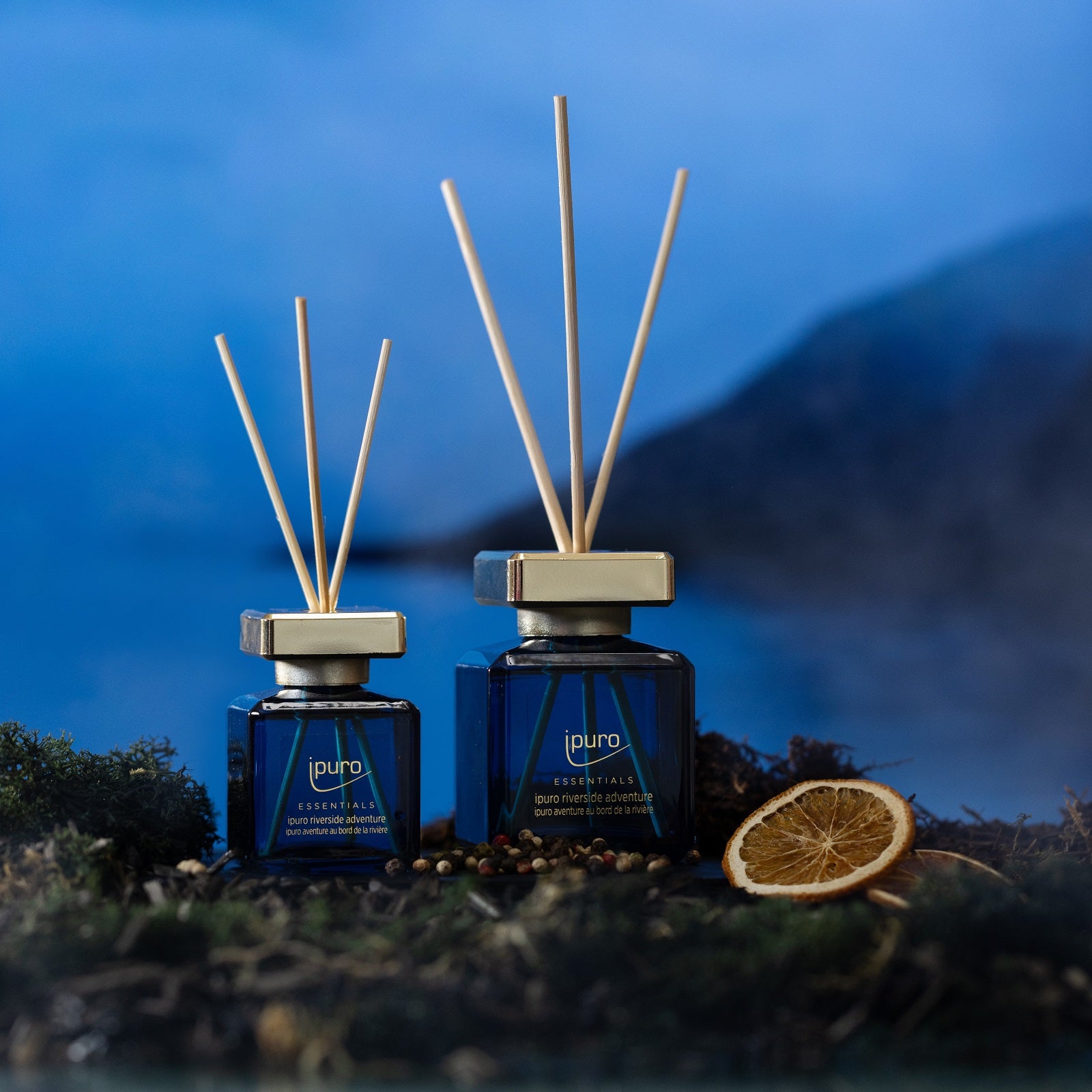 Zwei blaue ESSENTIALS ipuro riverside adventure Raumduft-Diffusoren von ipuro mit Holzstäben ruhen auf Moos neben einer getrockneten Orangenscheibe und verströmen einen frischen Duft vor einem verschwommenen, blauen See- und Berghintergrund.