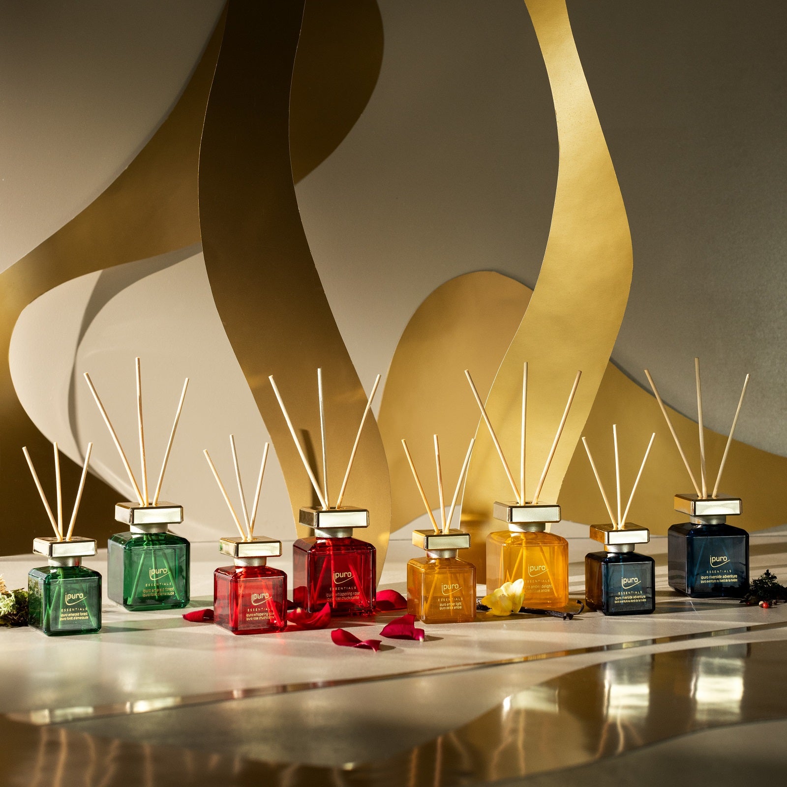 Ein Display mit acht leuchtenden ESSENTIALS ipuro whispering rose Raumduft-Tiegeln von ipuro mit Holzschilf und bunten Düften, die in einer Reihe auf einer reflektierenden Oberfläche vor einem abstrakten Hintergrund in Gold angeordnet sind.