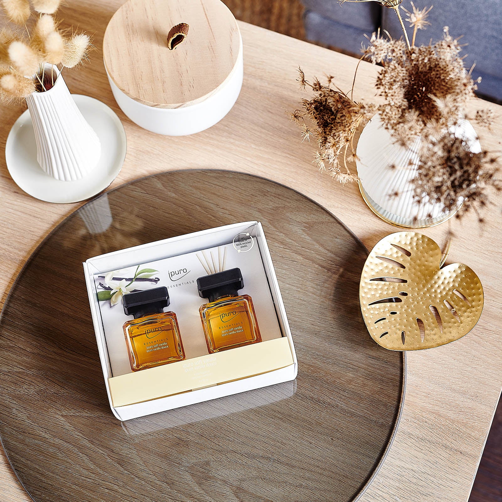 Auf einem Holztisch mit Glasplatte steht das ESSENTIALS ipuro soft vanilla Raumduft-Set von ipuro, bestehend aus zwei Duftflakons mit Vanille- und Sandelholznoten. Umgeben ist es von einer Trockenblumenvase und einer Schale in Form eines Blattgoldes.