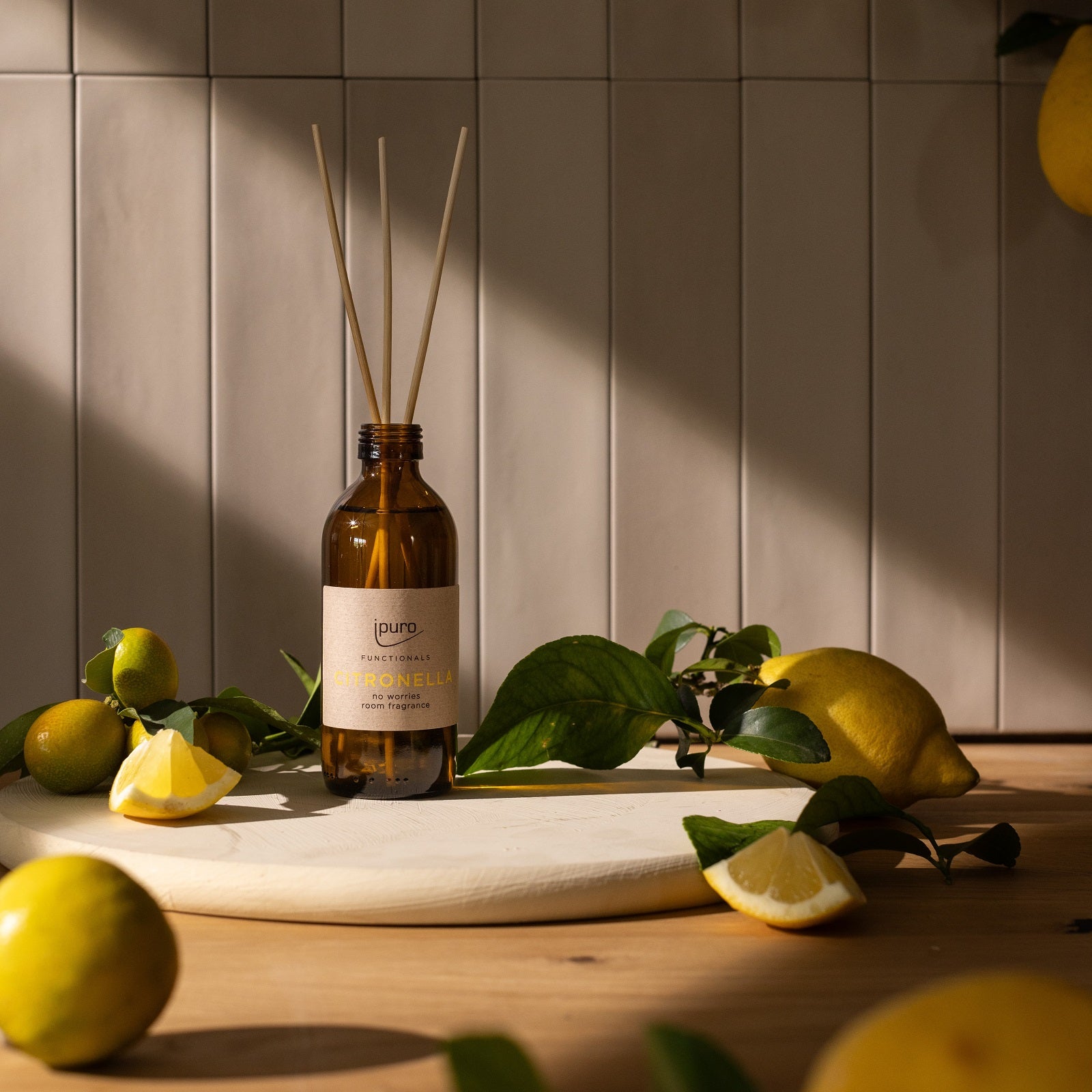 Ein FUNCTIONALS ipuro citronella Raumduft Diffusor von ipuro in einer braunen Glasflasche steht auf einer runden weißen Tafel zwischen frischen Zitronen, Zitronenscheiben und grünen Blättern mit natürlichem Licht und einer gefliesten Wand im Hintergrund.