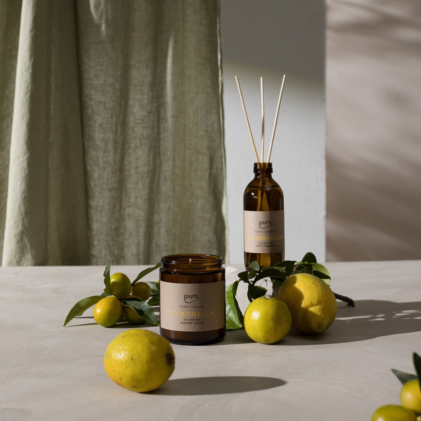 Der FUNCTIONALS ipuro citronella Raumduft von ipuro steht auf einem hellen Tisch mit leuchtenden Citronella-Etiketten, umgeben von Zitronen, Zitrusfrüchten und grünen Blättern. Ein grüner Vorhang und eine weiße Wand bilden einen weichen, ruhigen Hintergrund.