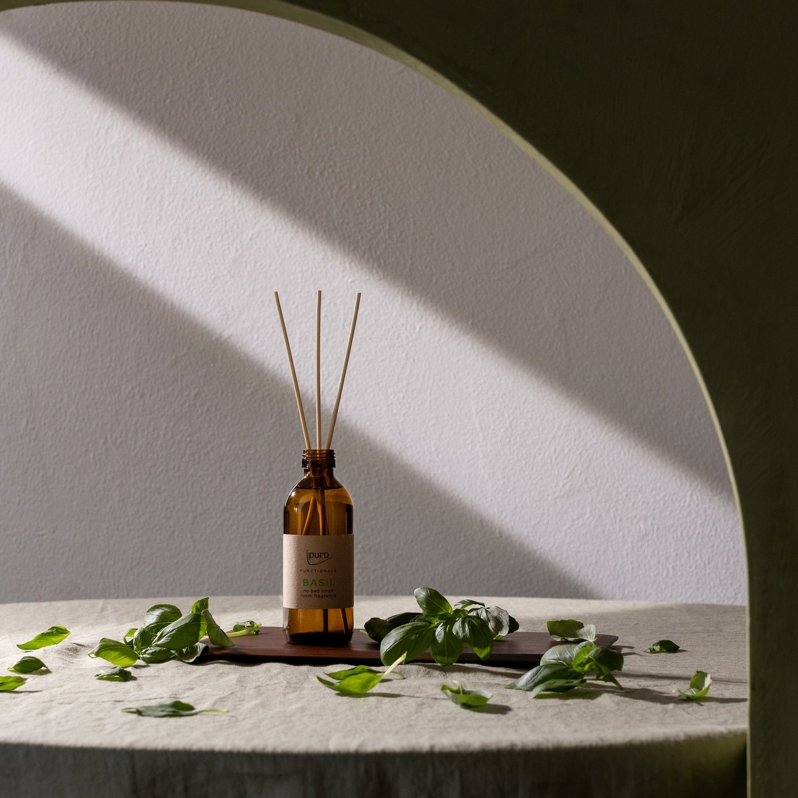 Ein brauner Glasdiffusor FUNCTIONALS ipuro basil Raumduft von ipuro aus Schilfrohr, beschriftet und umgeben von grünen Blättern, steht auf einem runden Tisch. Das Sonnenlicht strömt durch einen Torbogen und wirft Schatten auf die Wand und den Tisch.