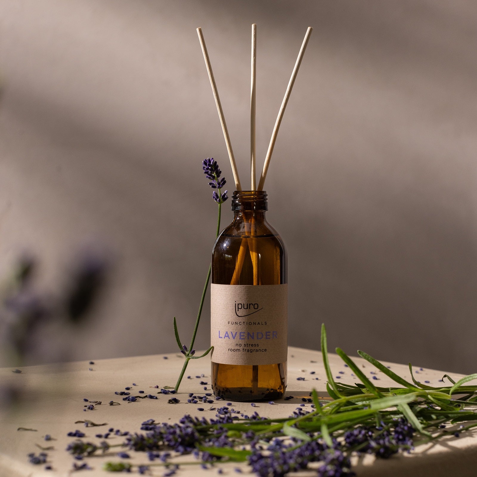 Eine braune Glasflasche mit der Aufschrift "ipuro FUNCTIONALS lavender Raumduft" von ipuro, mit Schilfrohrstäbchen und Lavendelzweigen, ruht auf einer beigen Fläche. Weiches natürliches Licht unterstreicht das entspannende Dufterlebnis.