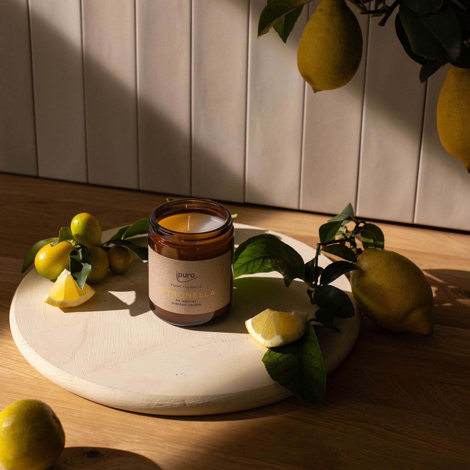 Die FUNCTIONALS ipuro citronella Duftkerze von ipuro aus Braunglas mit der Aufschrift Citronella verströmt einen erfrischenden Duft. Sie wird auf einem runden Holzbrett mit Zitronen, Zitronenscheiben, grünen Blättern und Zitrusfrüchten im Sonnenlicht präsentiert.