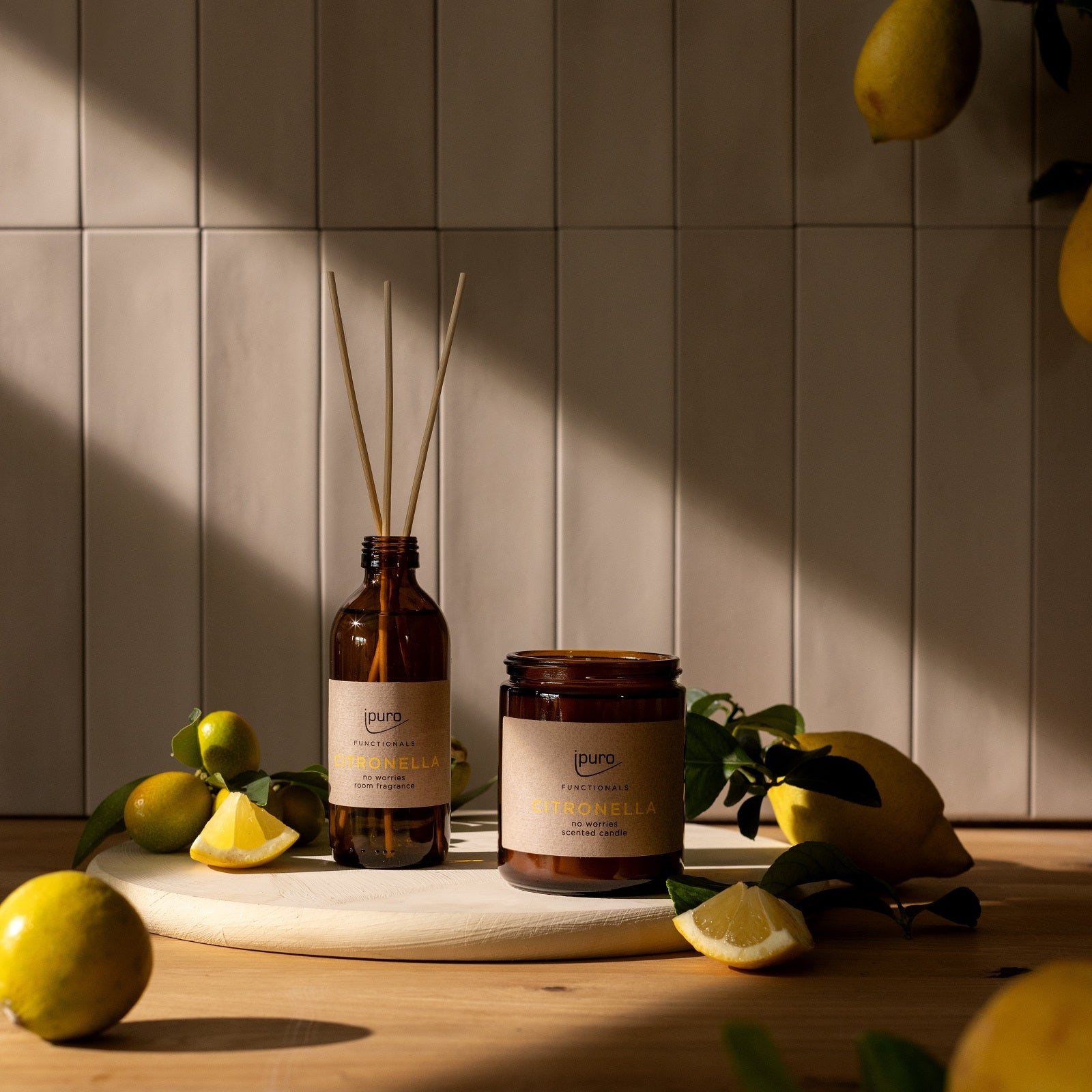 Eine ipuro FUNCTIONALS Citronella-Duftkerze steht auf einem weißen Tablett mit Zitronen, Limetten und Blättern, arrangiert auf Holz. Das Sonnenlicht wirft Schatten auf die geflieste Wand dahinter.