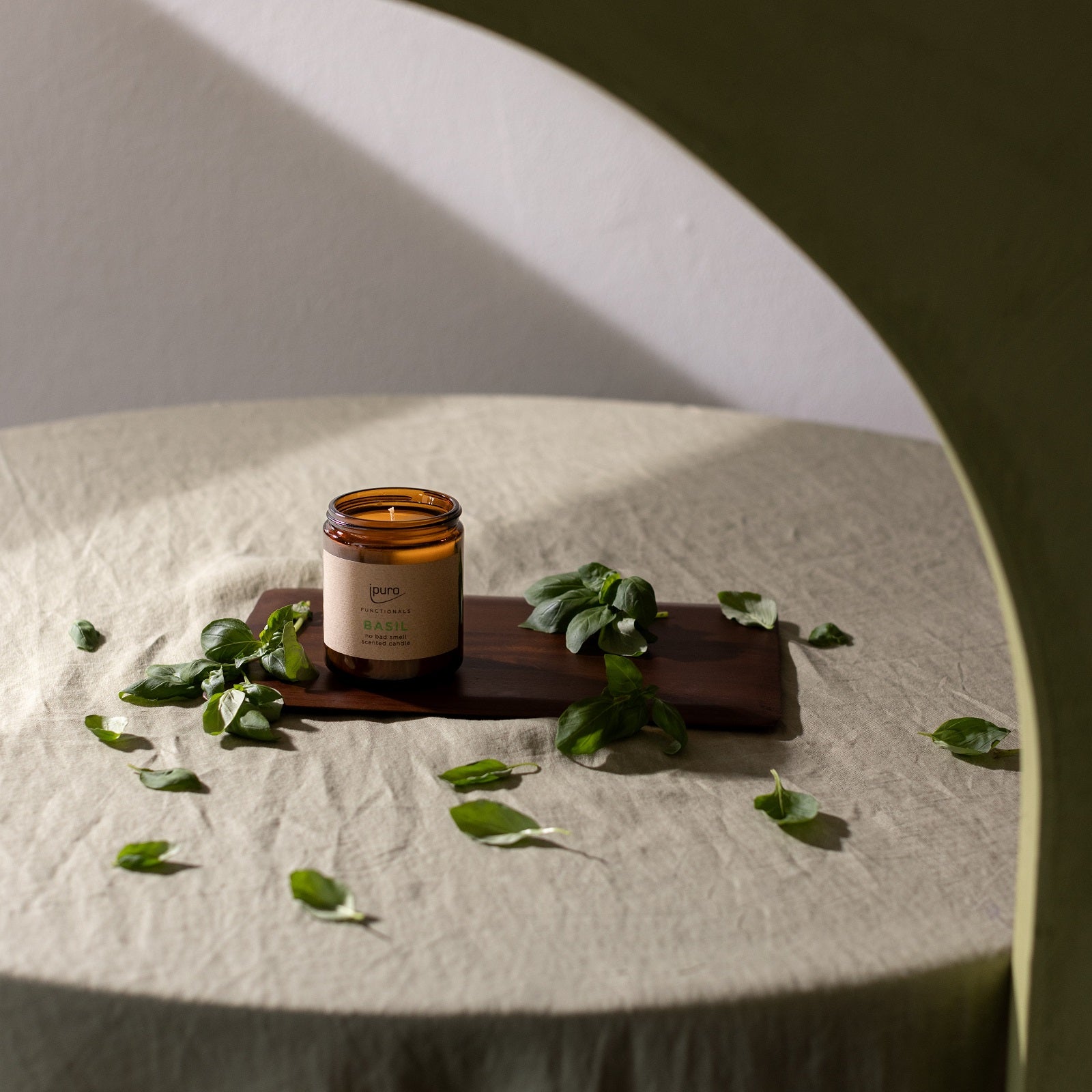 Ein braunes Glasgefäß der FUNCTIONALS ipuro basil Duftkerze von ipuro steht auf einem Holztablett auf einem beigen Stofftisch, umgeben von frischen grünen Basilikumblättern und einer bogenförmigen grünen Struktur, die die Küchenduft-Szene einrahmt.
