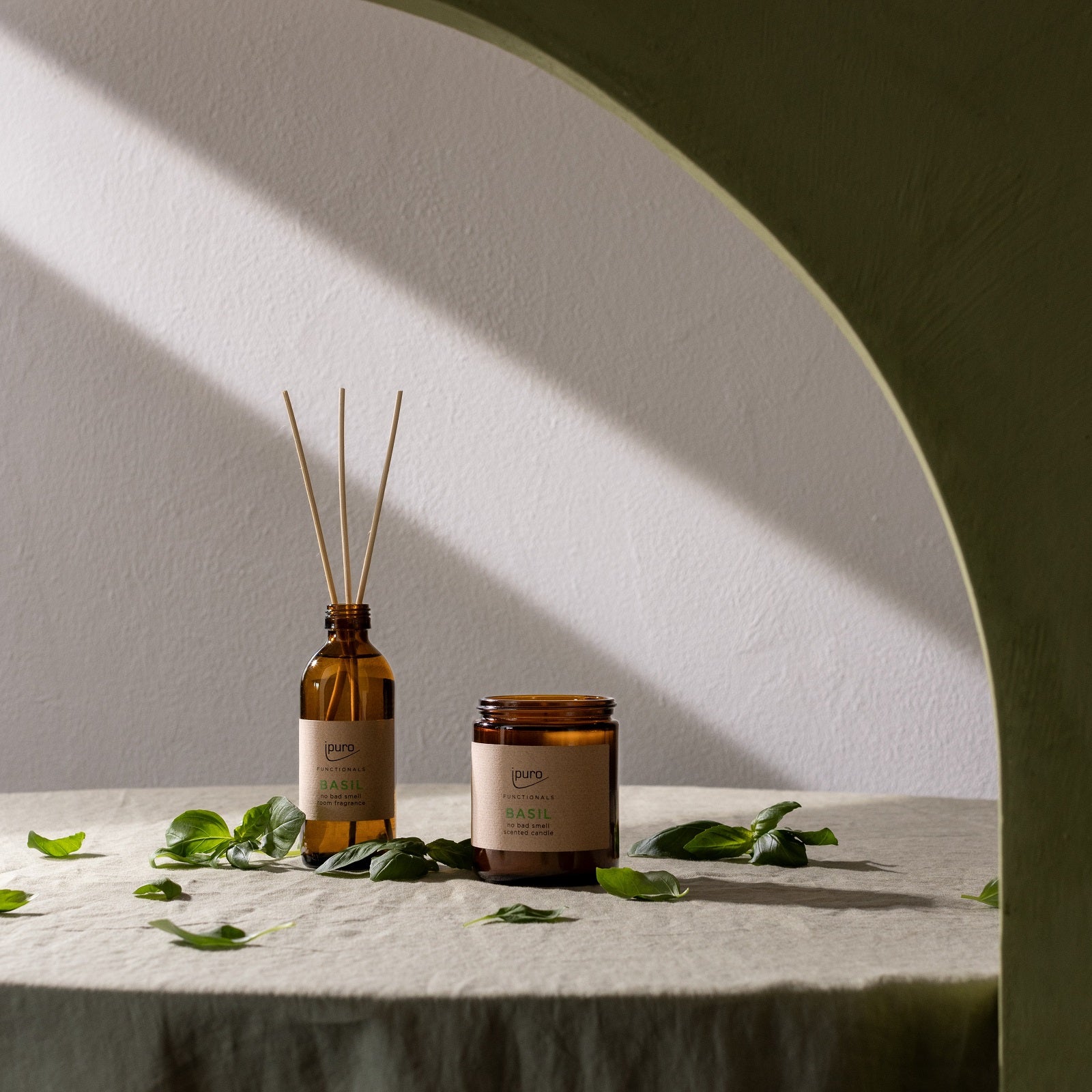 Die FUNCTIONALS ipuro basil Duftkerze und der Braunglasdiffusor von ipuro, besetzt mit frischen Basilikumblättern auf einem leichten Tuch, werden umrahmt von einem grünen Bogen in sanfter Naturlicht-Küchenduft powered by Dufttechnologie gegen Gerüche.