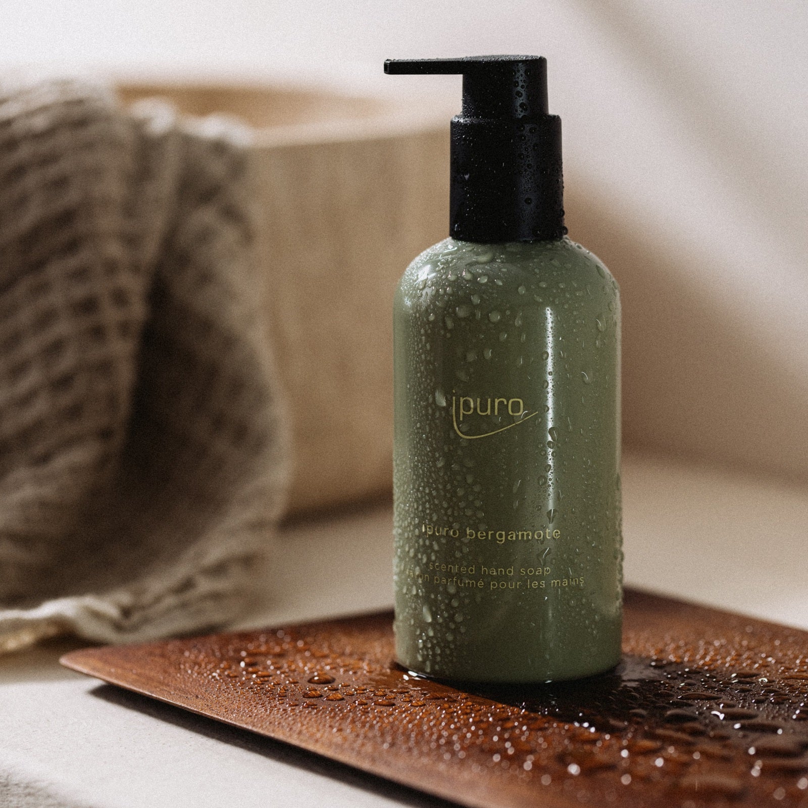 Eine grüne Pumpflasche ipuro CLASSIC bergamote Handseife steht auf einem braunen Tablett mit Wassertropfen; im unscharfen Hintergrund sorgen ein strukturiertes beiges Tuch und eine Holzbox für einen Hauch asiatischer Frische.