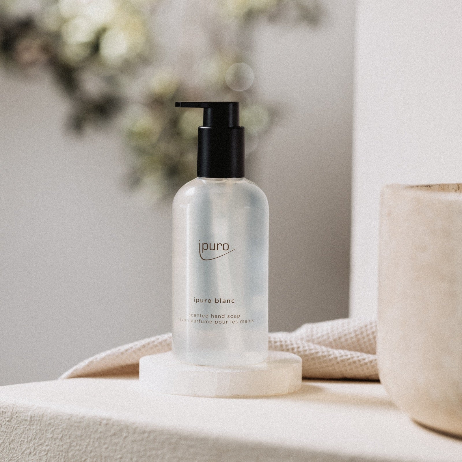 Eine transparente Flasche CLASSIC ipuro blanc Handseife von ipuro mit einer schwarzen Pumpe ruht auf einem runden weißen Untersetzer auf beigem Stoff und erfüllt die Luft mit Noten von Minze und Sandelholz vor einem weichen, neutralen Hintergrund.