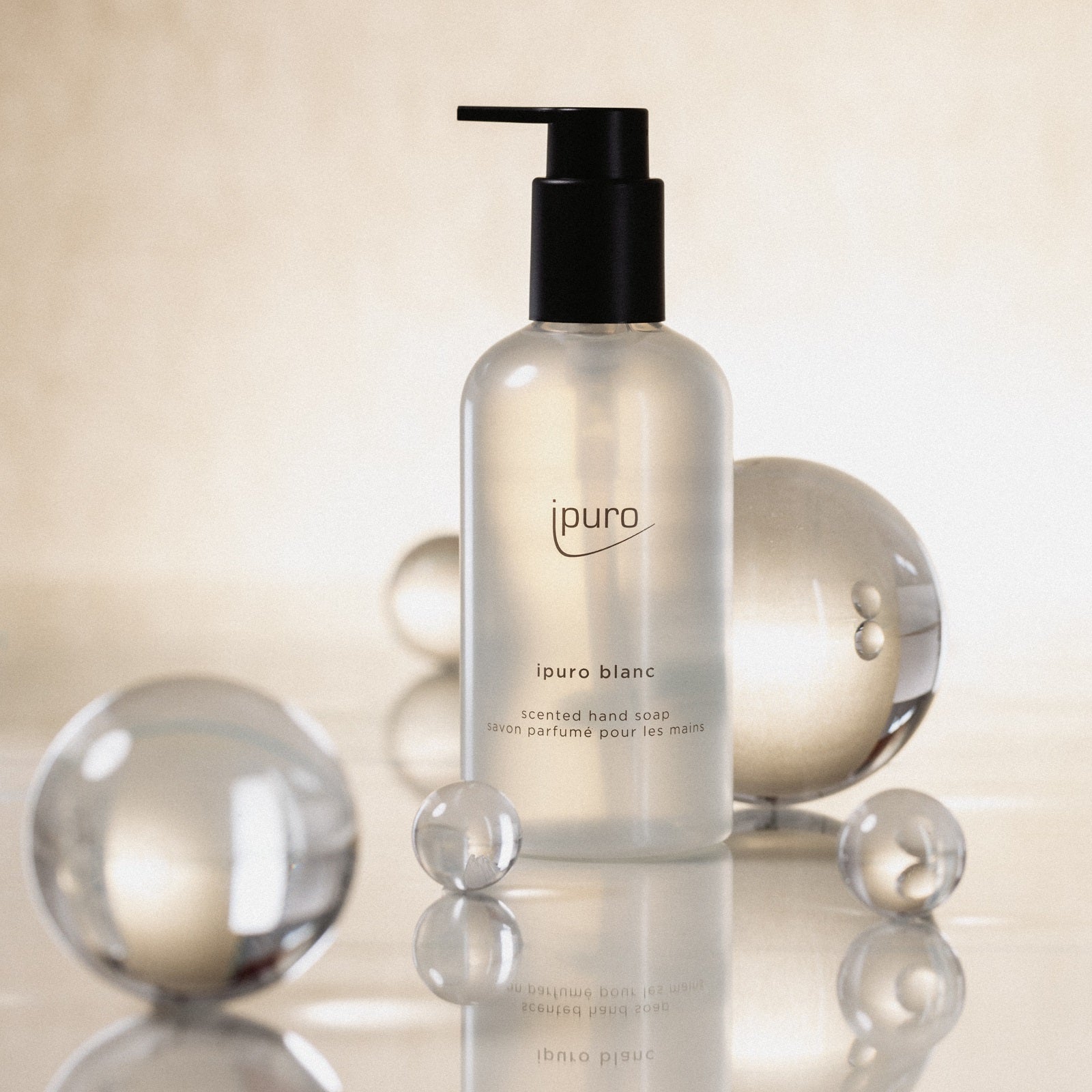 Eine aufrecht stehende CLASSIC ipuro blanc Handseife-Pumpenflasche von ipuro mit Sandelholz-Noten wird zwischen reflektierenden Glaskugeln auf einem sanft strukturierten, hellen Hintergrund präsentiert, wodurch ein eleganter, minimalistischer Look entsteht.