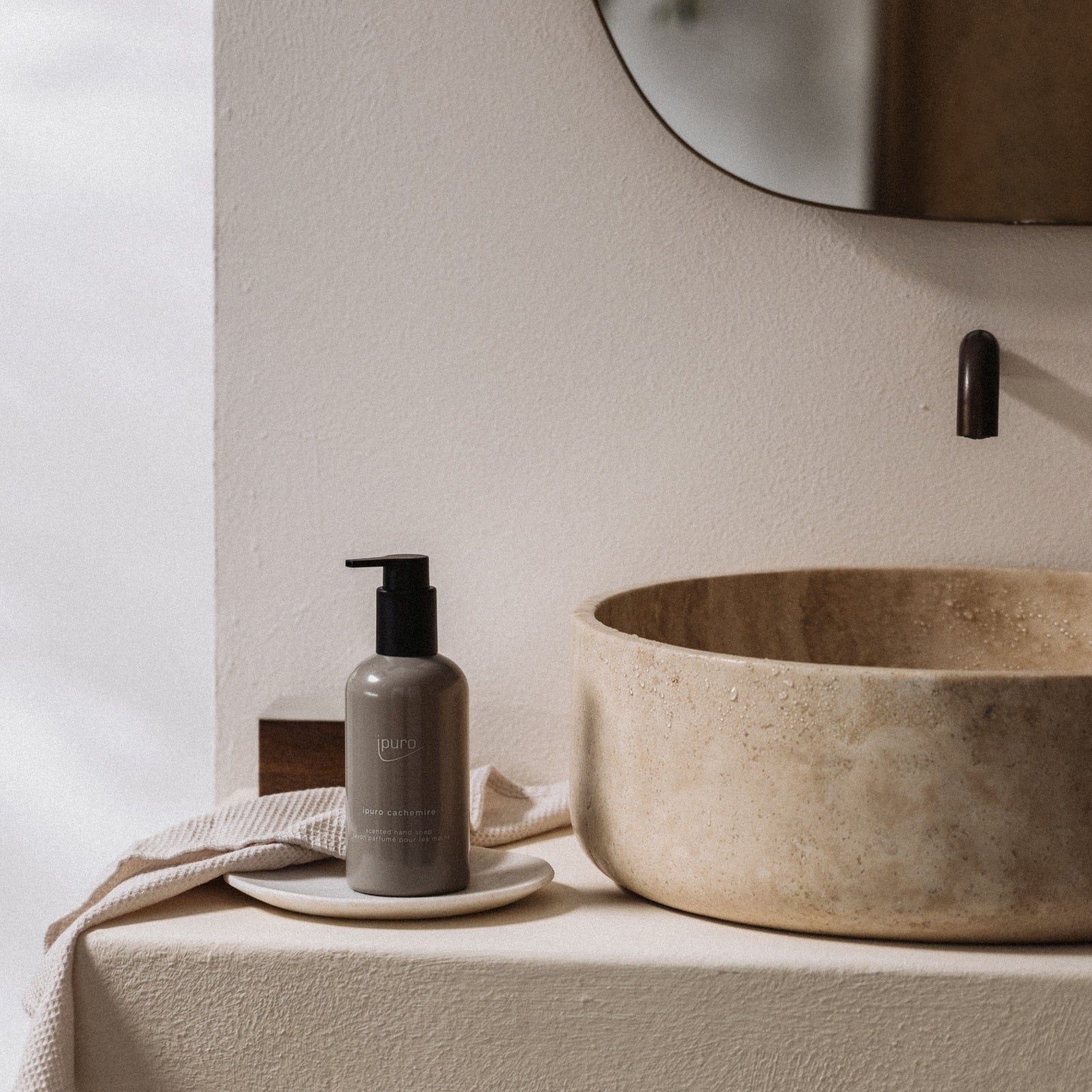 Eine minimalistische Badezimmerszene mit einem runden Steinwaschbecken, einem Wandwasserhahn, einem beigen Handtuch und einem grauen ipuro CLASSIC cachemire Handseifenspender auf einer kleinen weißen Schale neben dem Waschbecken. Darüber ist teilweise ein Spiegel zu sehen.