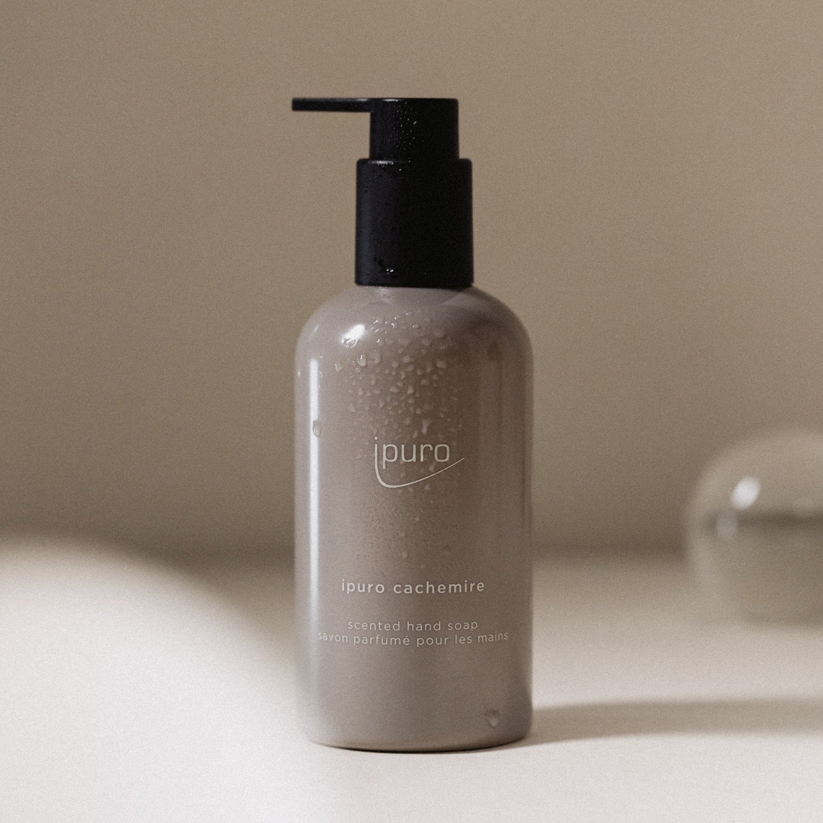 Ein beiger Flakon von CLASSIC ipuro cachemire Handseife von ipuro, beduftet mit Zedernholz und einem Hauch von Rose, steht auf einer neutralen Fläche mit Wassertropfen; der sanft verschwommene Hintergrund verleiht Eleganz.