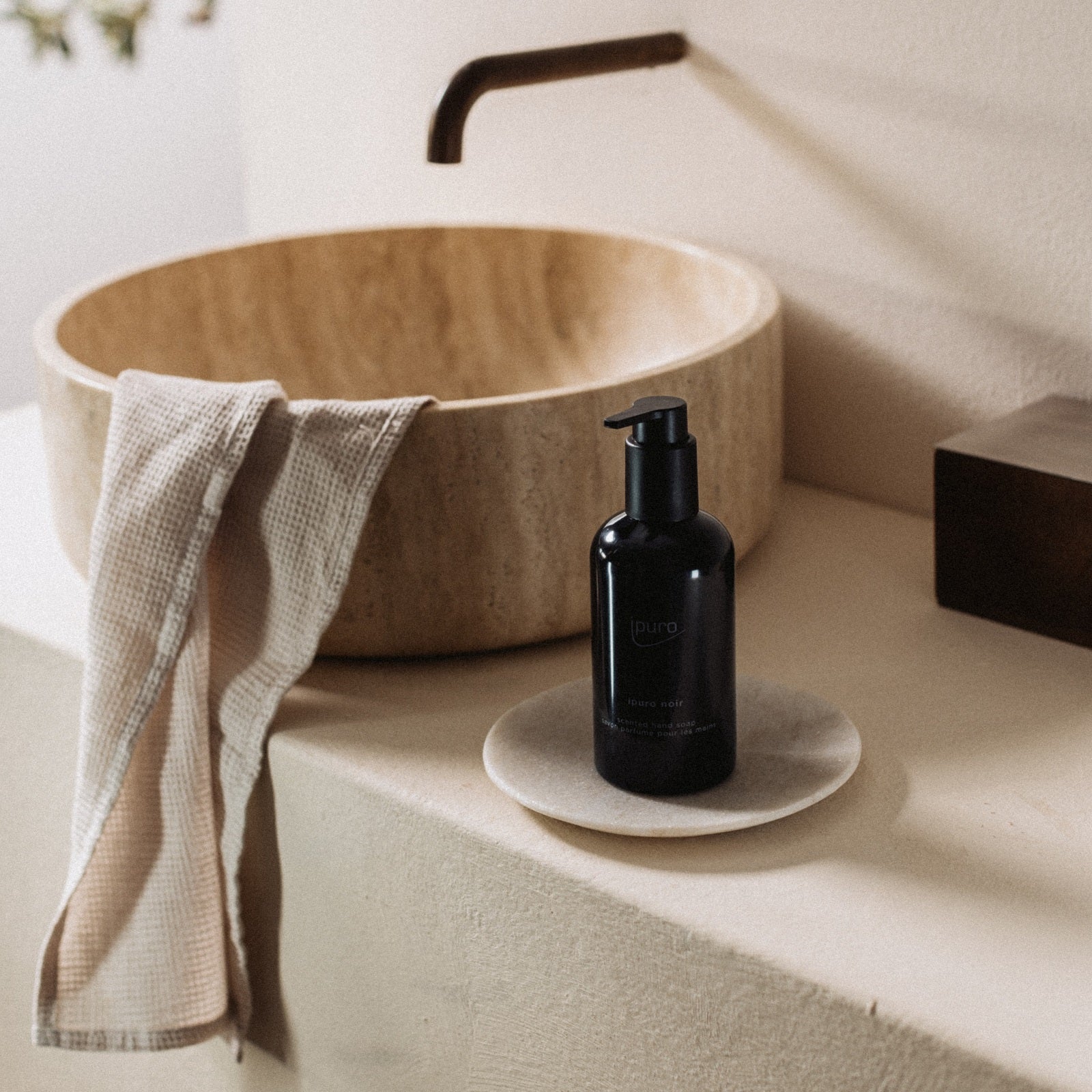 Ein beiges Steinwaschbecken mit bronzefarbenem Wasserhahn, ein strukturiertes Handtuch und der schwarze Seifenspender CLASSIC ipuro noir Handseife von ipuro auf einer weißen Schale stehen auf einer cremefarbenen Arbeitsplatte und erinnern an die Wärme von Sandelholz und orientalischem Parfum.