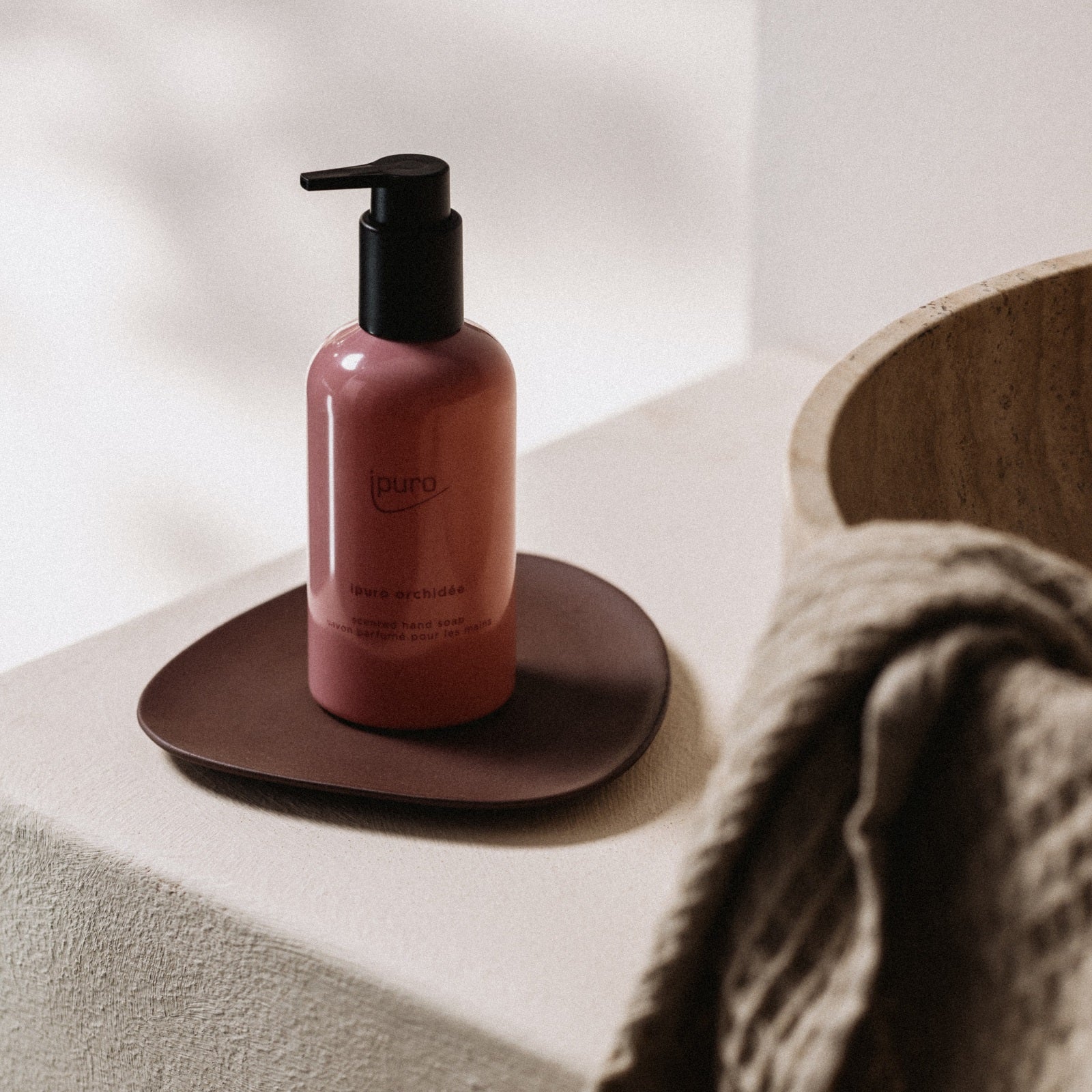 Ein rosafarbener ipuro CLASSIC orchidée Handseife-Flakon mit schwarzer Pumpe steht auf einer braunen Schale neben einem beigen Handtuch und einer Holzschale, die auf einer hellen Fläche angeordnet sind. Der Duft von sizilianischer Limone trägt zum beruhigenden Ambiente bei.
